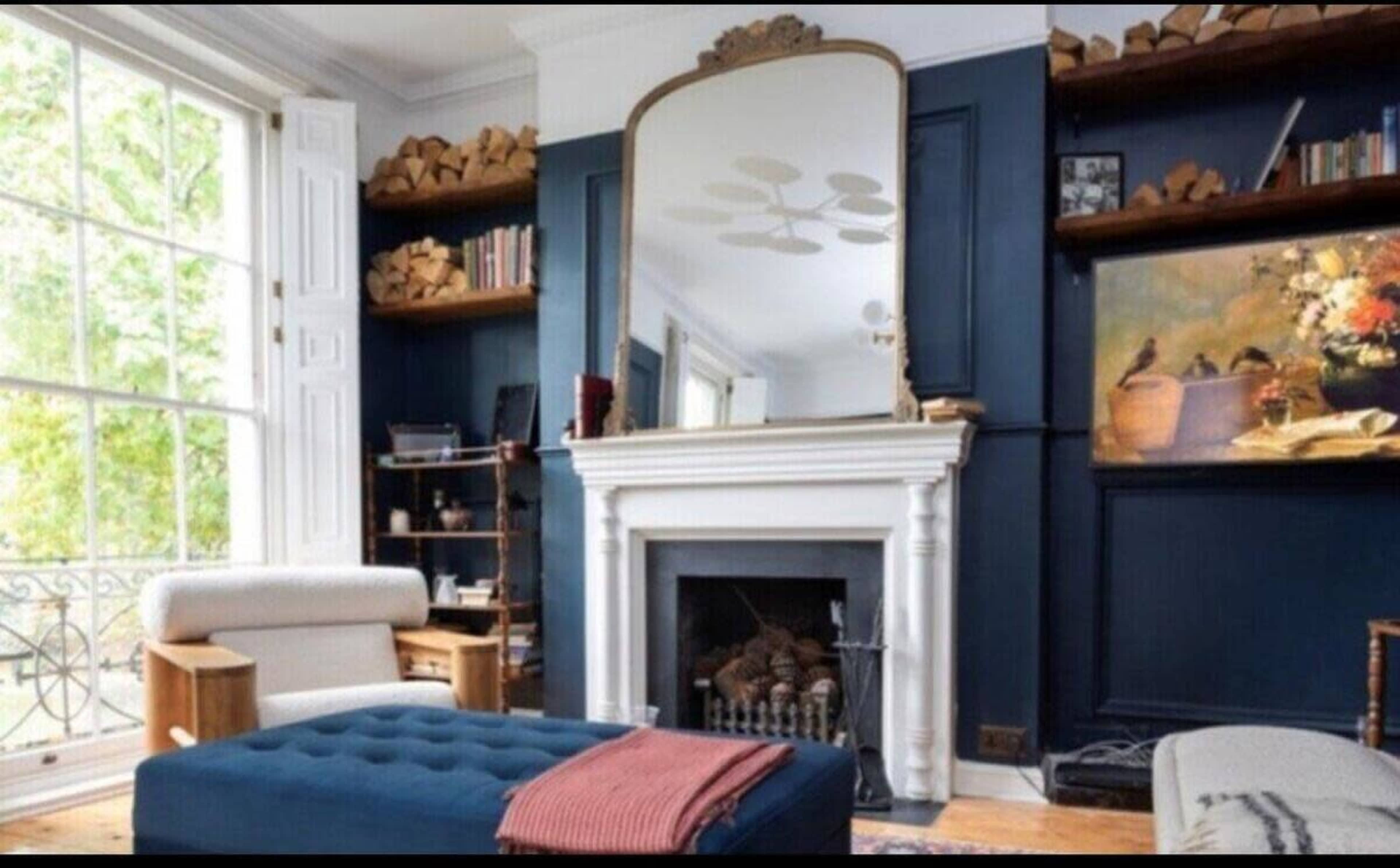 A stylish living room featuring a blue accent wall, a white fireplace with a mirror above it, a large ottoman, and wooden shelves with books and logs.