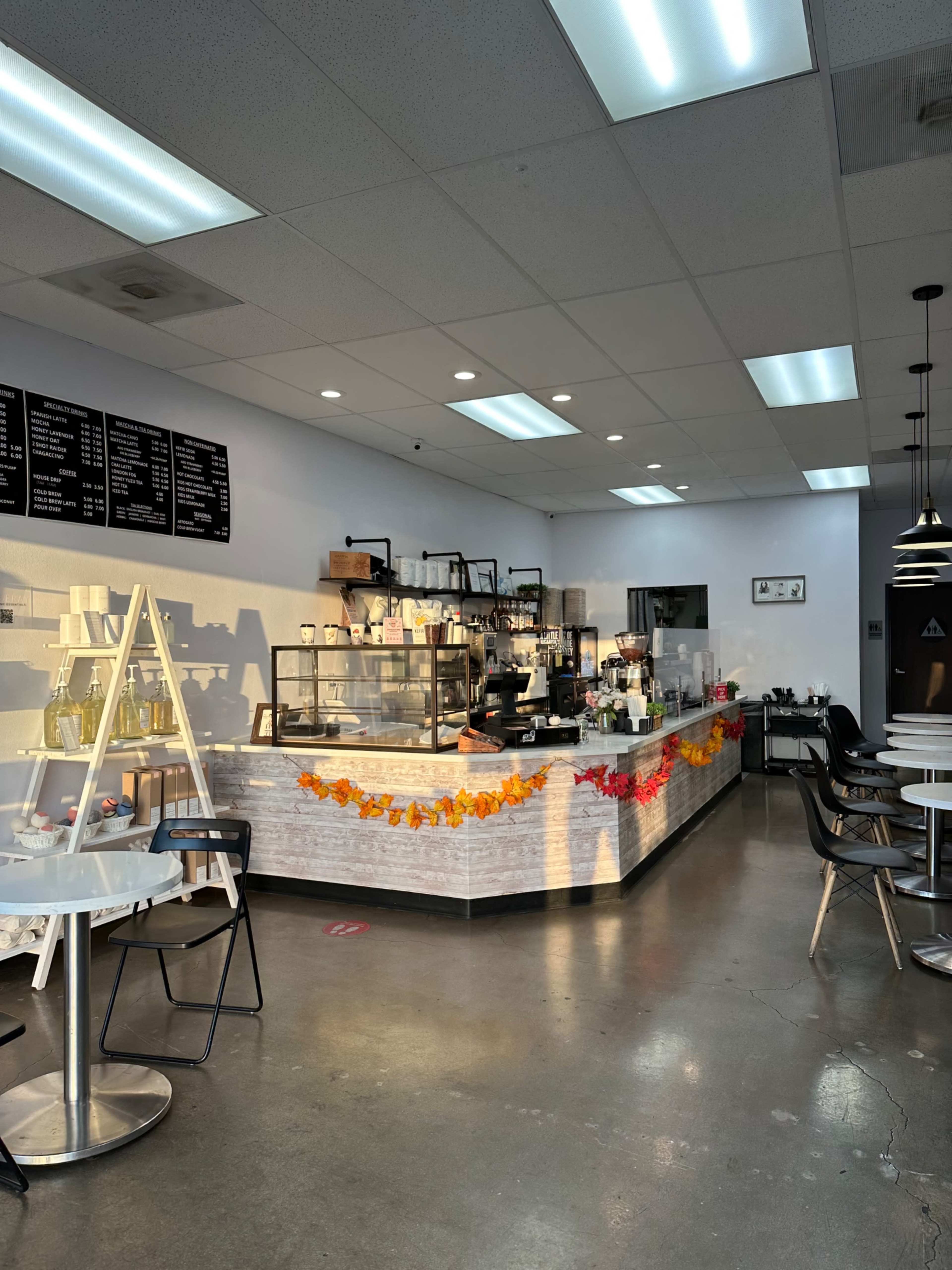 The interior of a café features a service counter with an assortment of baked goods and beverages, surrounded by tables and chairs in a brightly lit space.