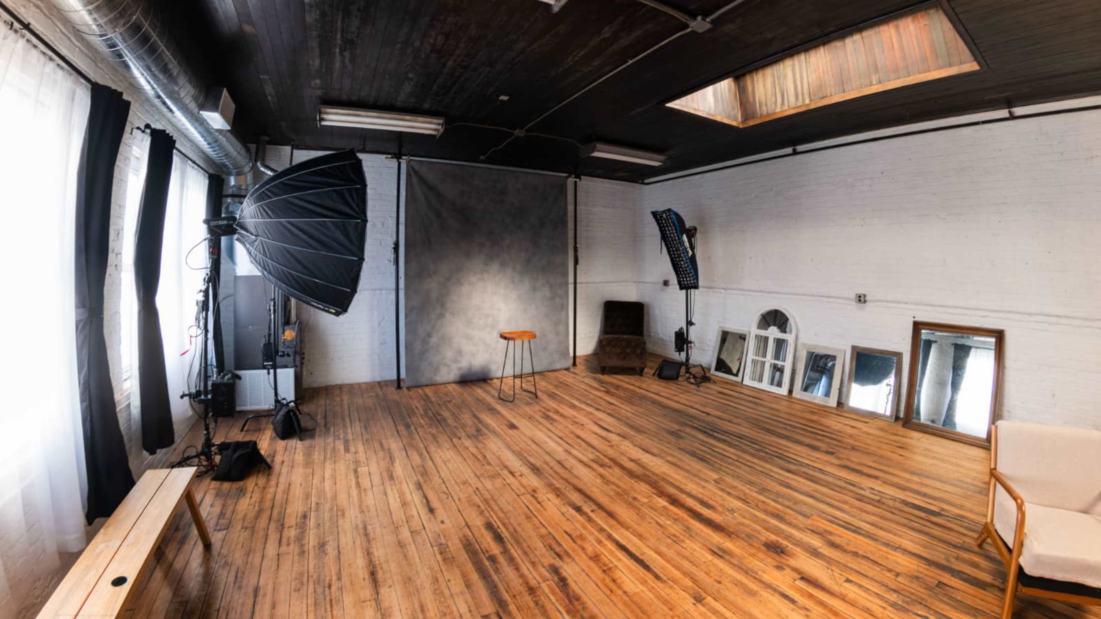 The image depicts a spacious photography studio featuring wooden floors, a backdrop, lighting equipment, and various furniture.