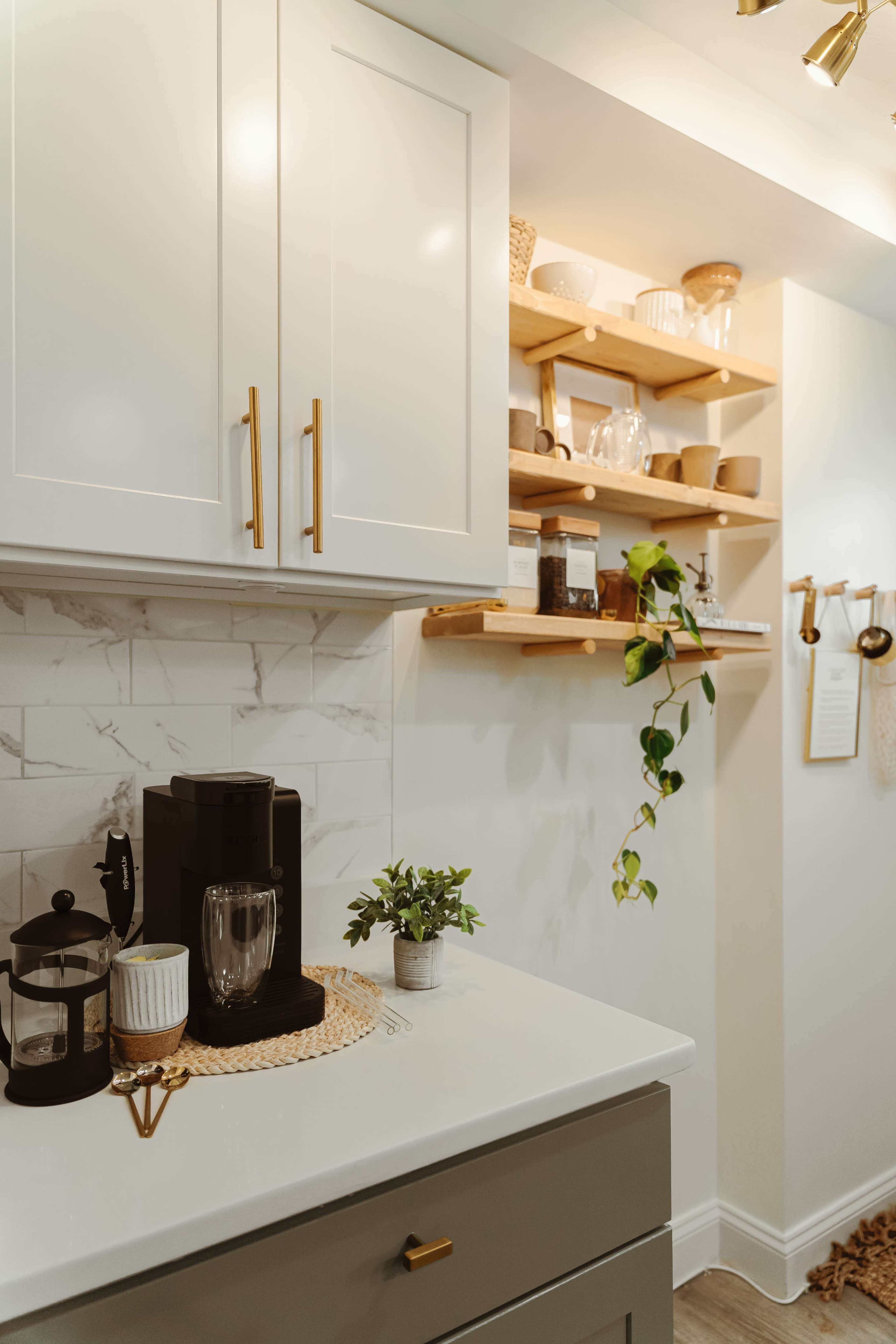 The image shows a kitchen corner featuring a coffee maker on a countertop, white cabinets with gold handles, and wooden shelves displaying various kitchen items and a potted plant.