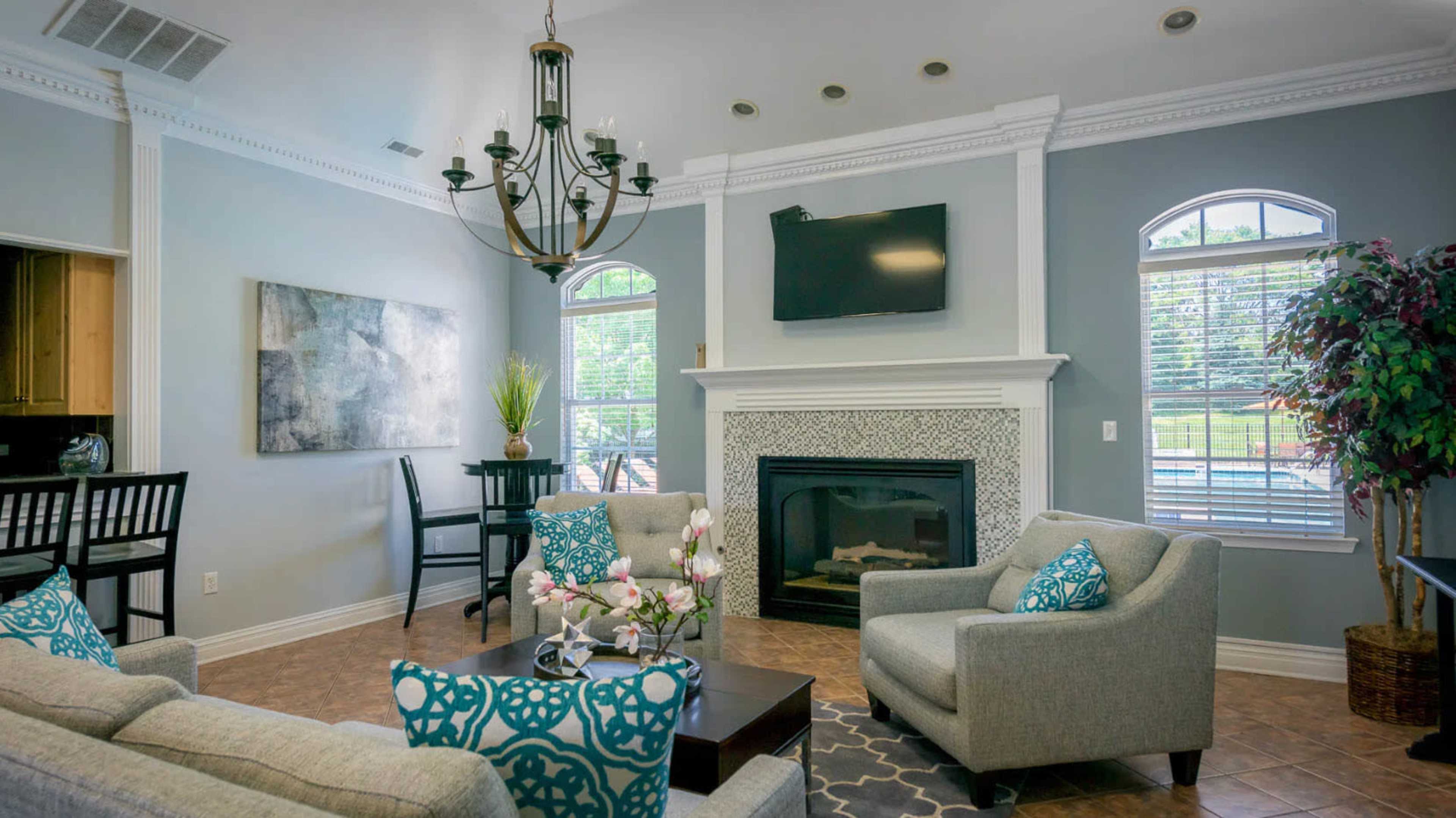 The image shows a living room with a chandelier, a fireplace with a patterned tile surround, and a seating area featuring gray sofas adorned with blue cushions.