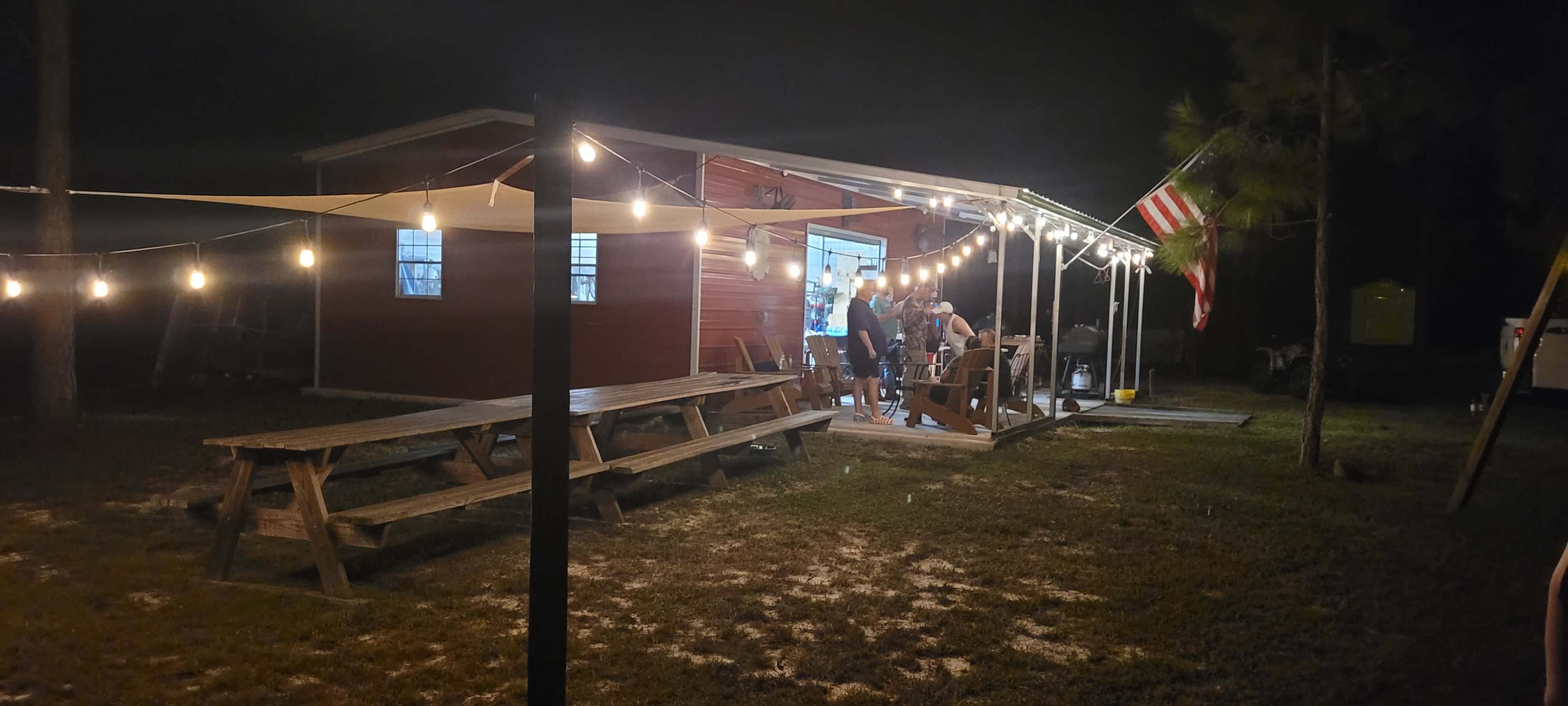 A group of people gathers on a porch of a red building at night, with string lights and an American flag hanging nearby.