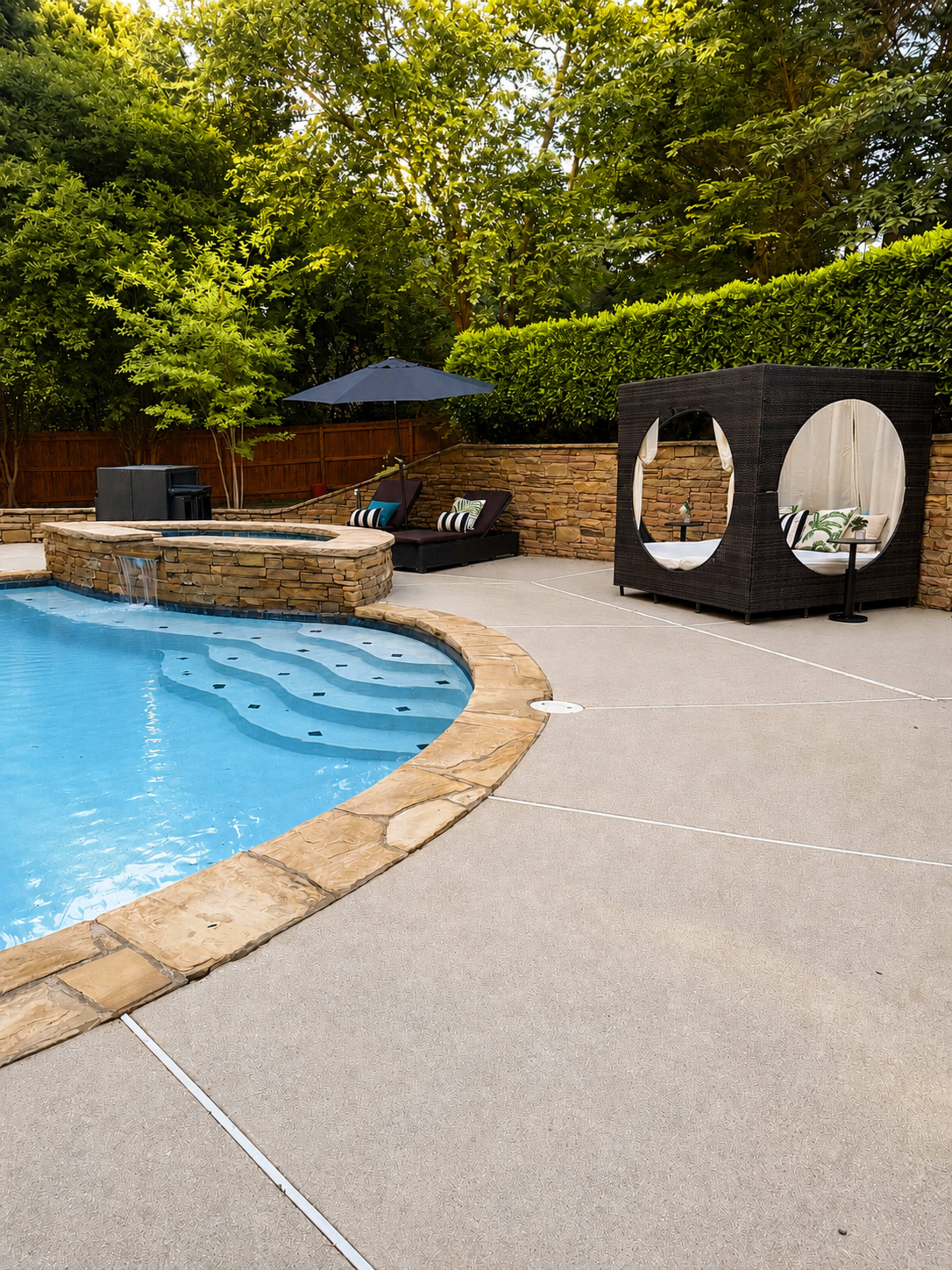 The image shows a backyard pool area featuring a stone-lined pool, a lounge chair with an umbrella, and a covered seating area next to a green hedge.