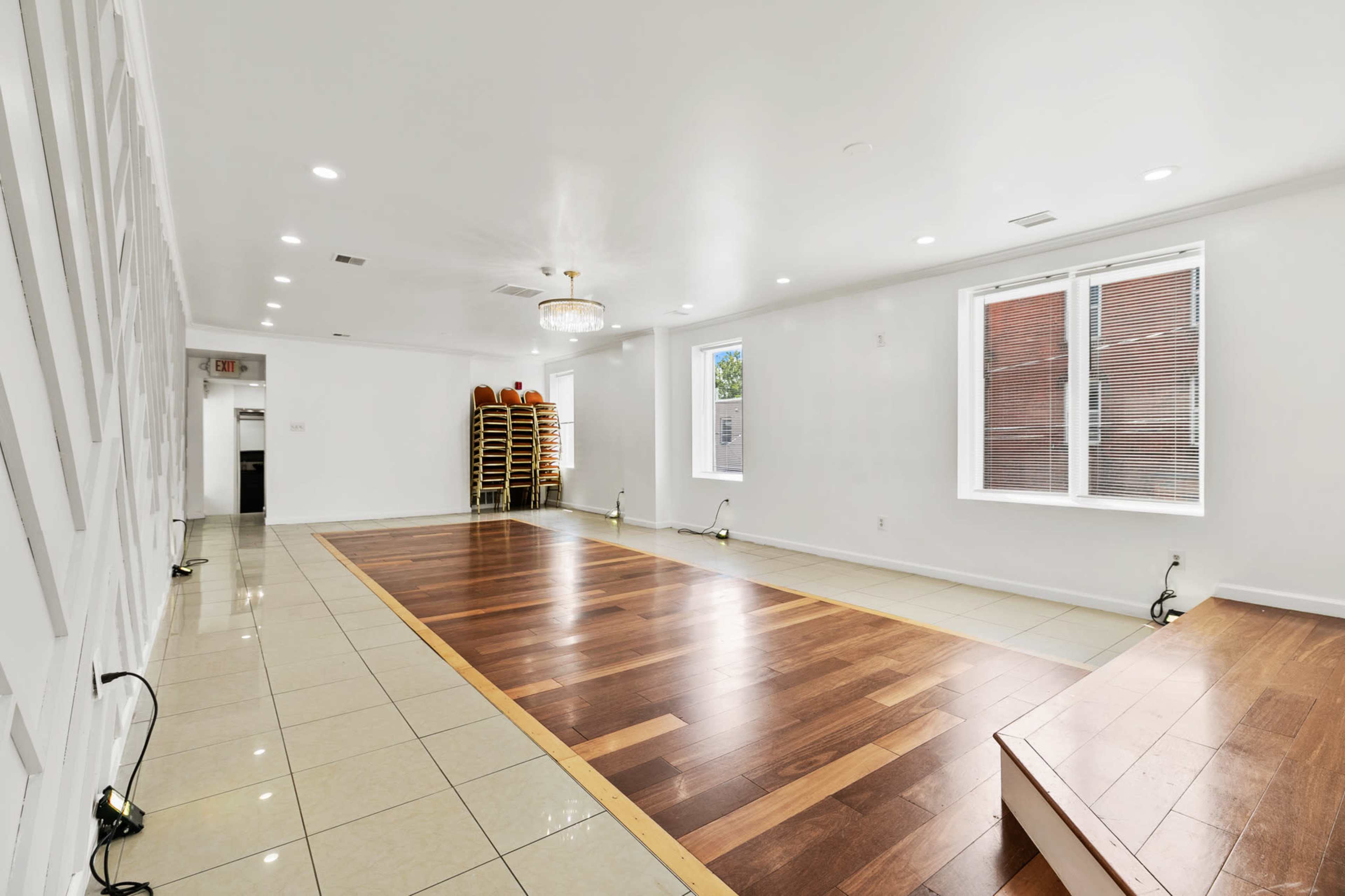 The image shows a brightly lit empty room with a combination of tiled and wooden flooring, featuring large windows and a row of stacked chairs against the wall.