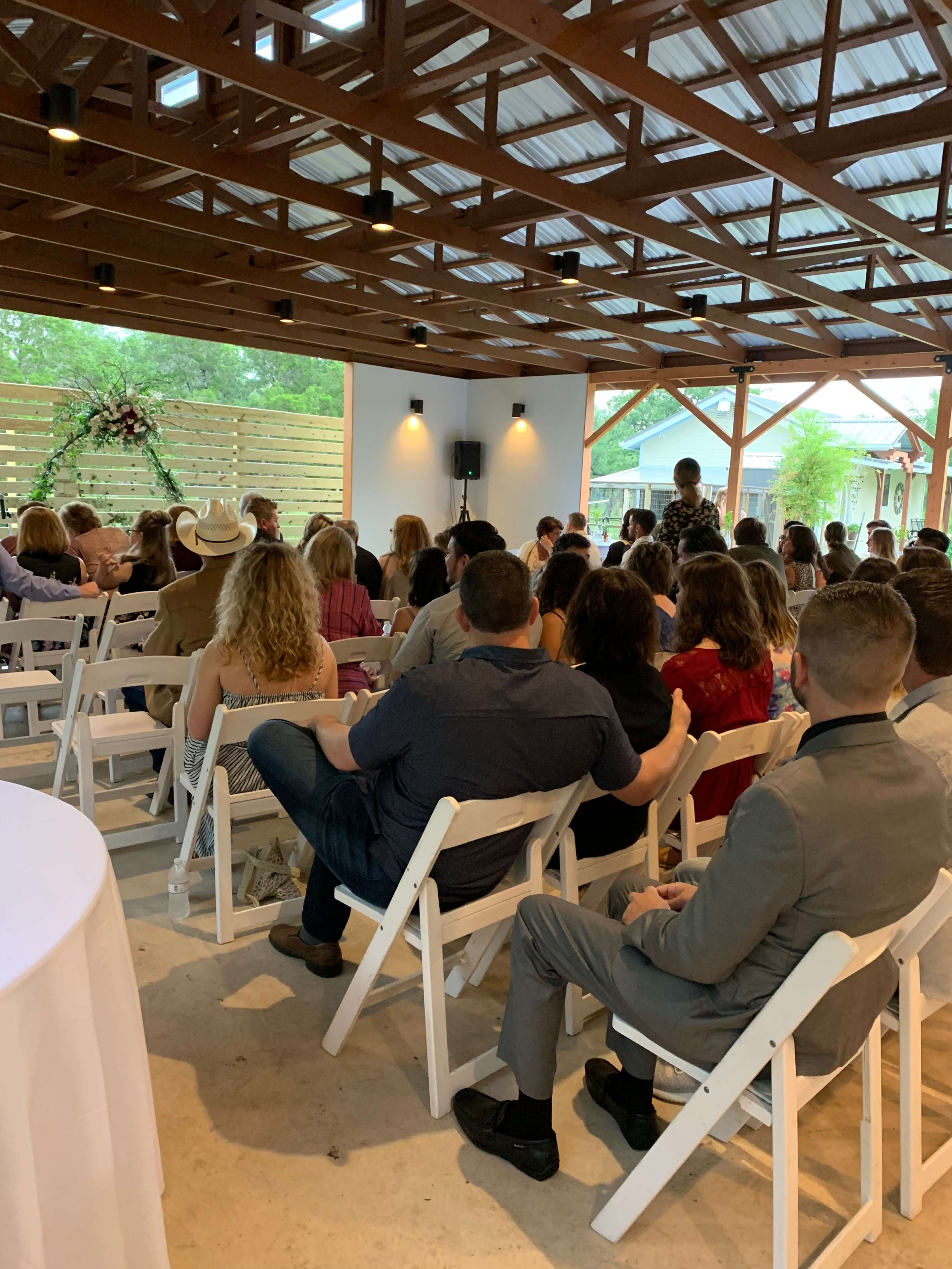 A group of people is seated in rows on white folding chairs in a spacious indoor venue with wooden beams and large windows.