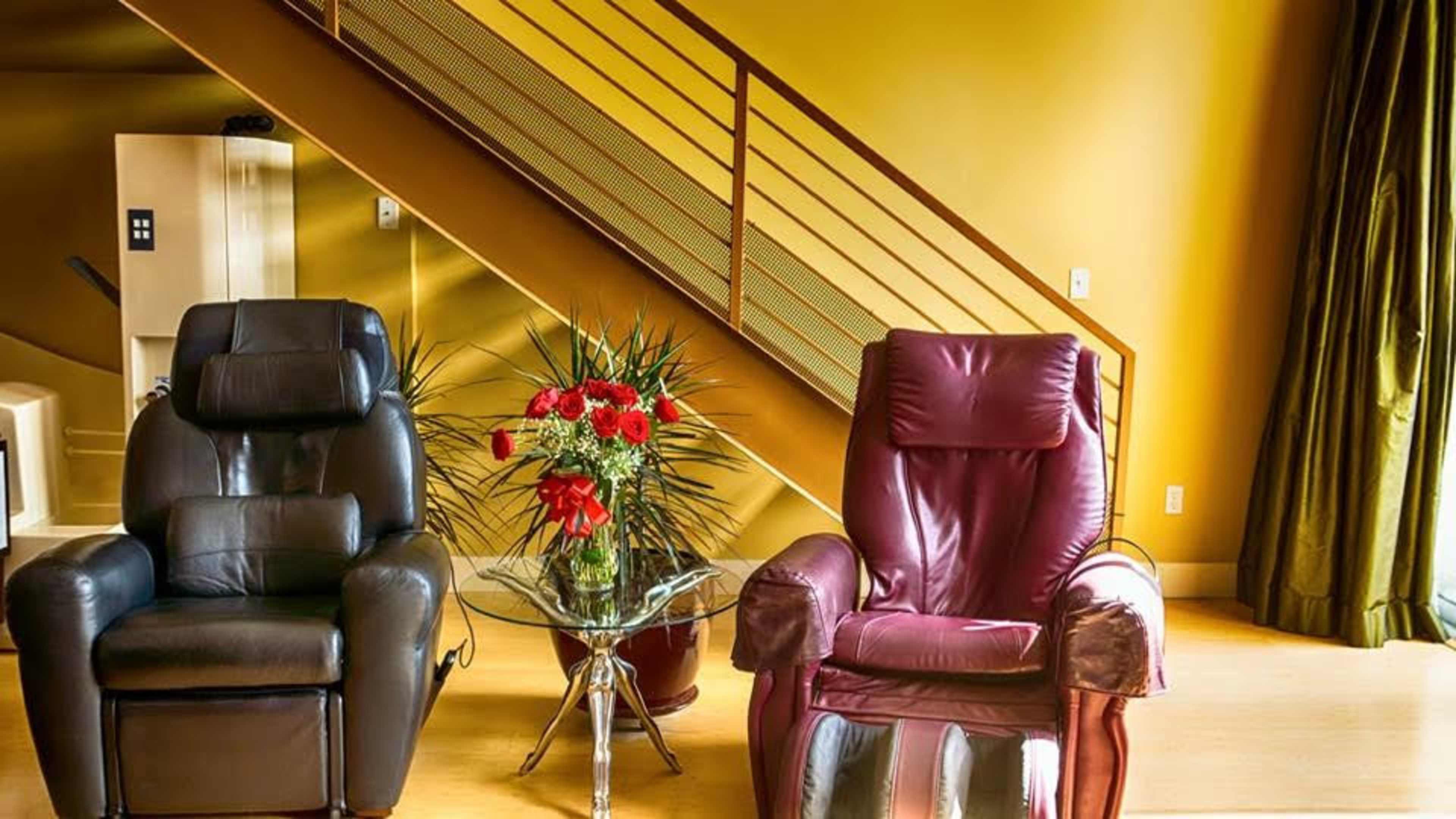 Two recliner chairs, one black and one burgundy, positioned near a small table with a bouquet of flowers, against a backdrop of a yellow wall and a staircase.