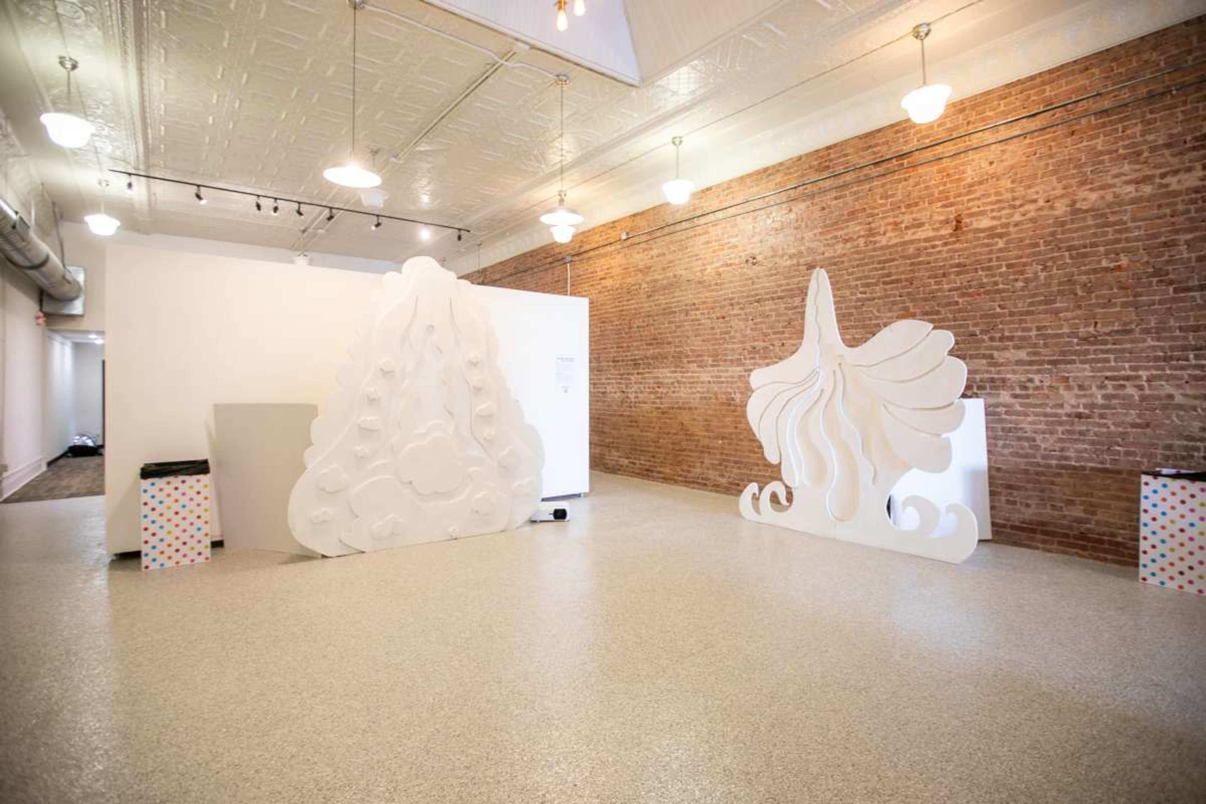 The image shows an art gallery featuring two large, white sculptural installations against a backdrop of exposed brick walls.
