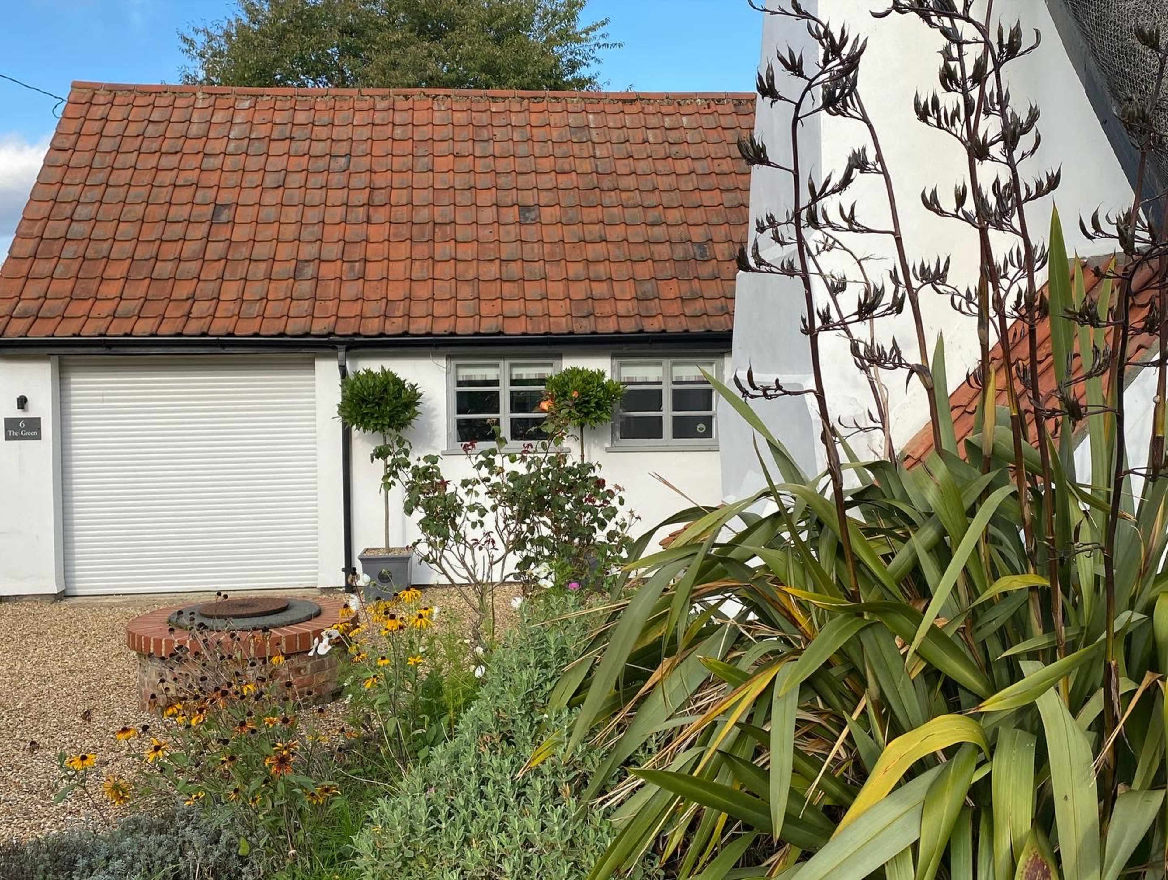 A white house with a red-tiled roof features a garage door and is surrounded by a garden with flowers and green shrubs.