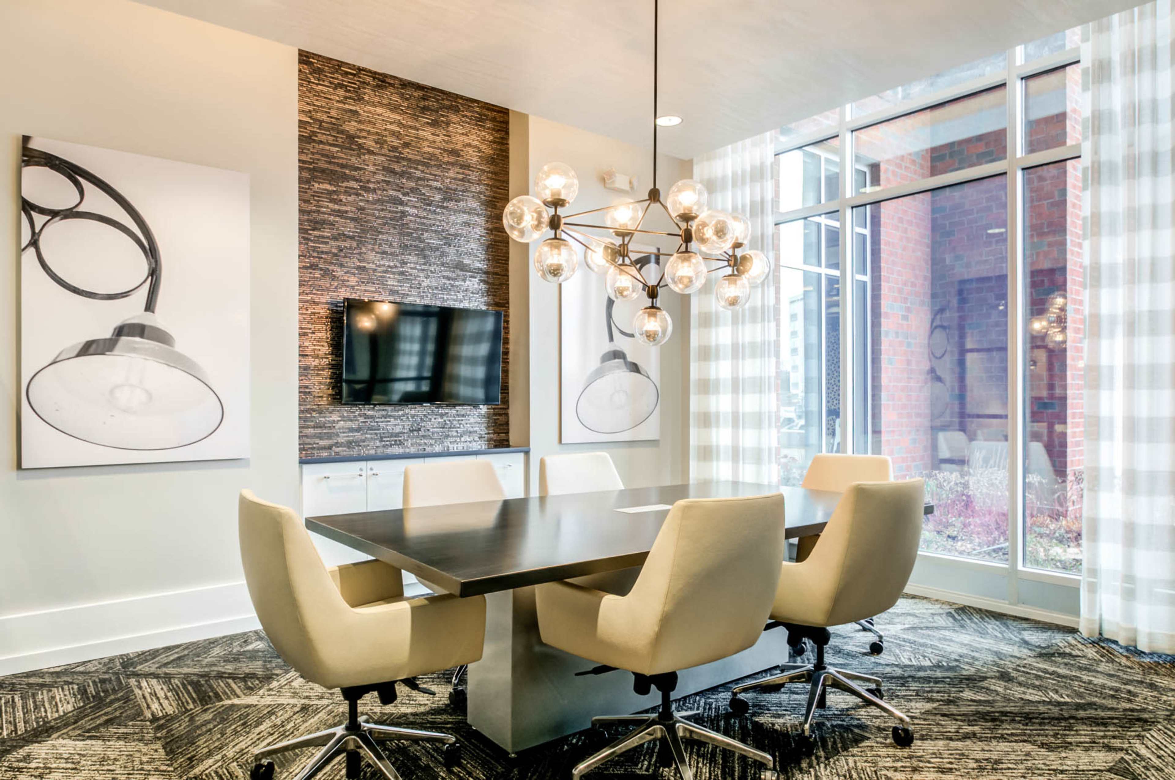 A modern conference room features a large table surrounded by cream-colored chairs, with a chandelier above and a wall-mounted television.