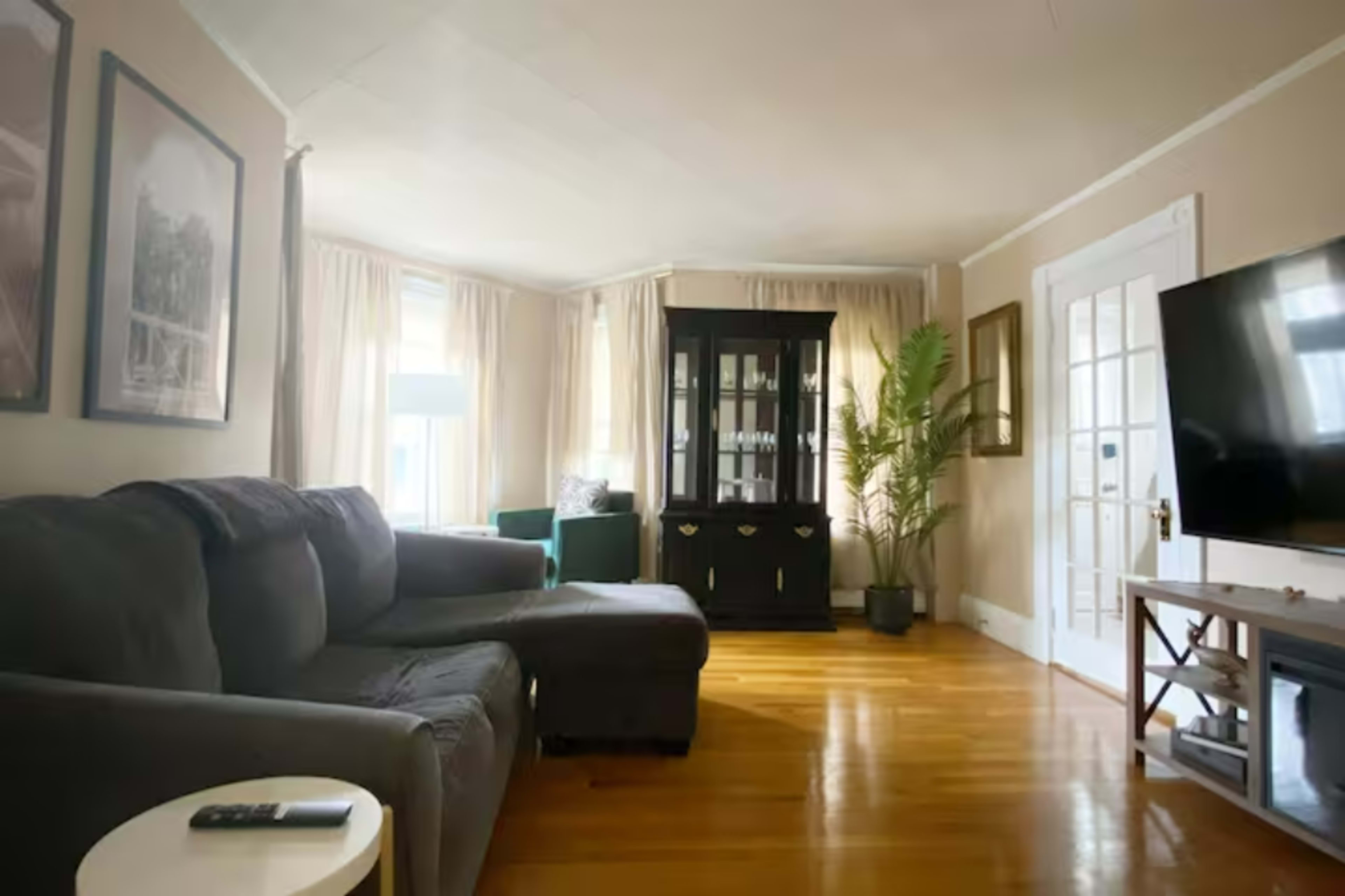 A cozy living room featuring a dark sofa, a small white side table, a black cabinet, and a television on a wooden stand, with large windows dressed in light curtains.