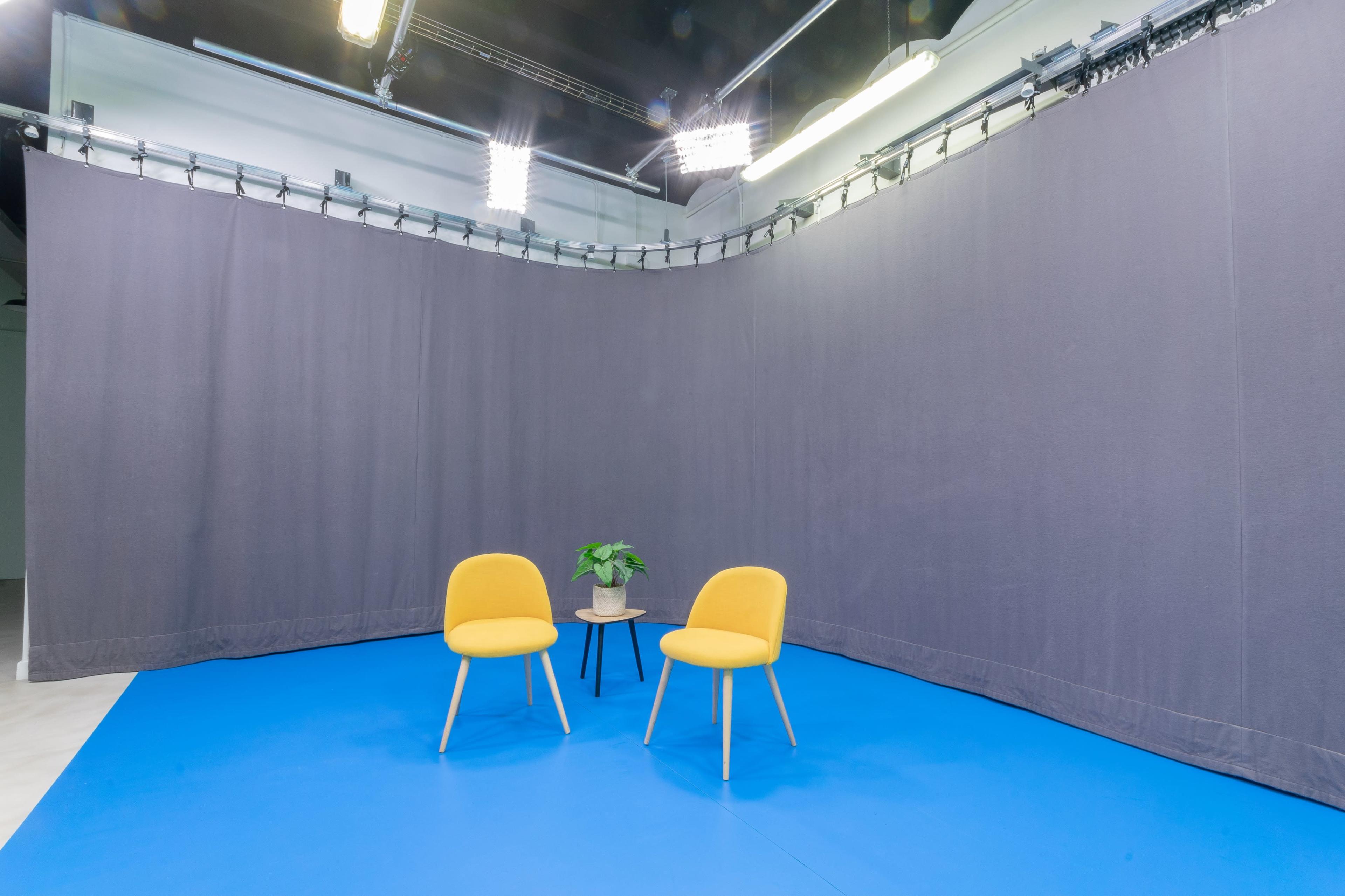 A studio setup features two yellow chairs and a small potted plant on a table against a backdrop of gray curtains and a blue floor.