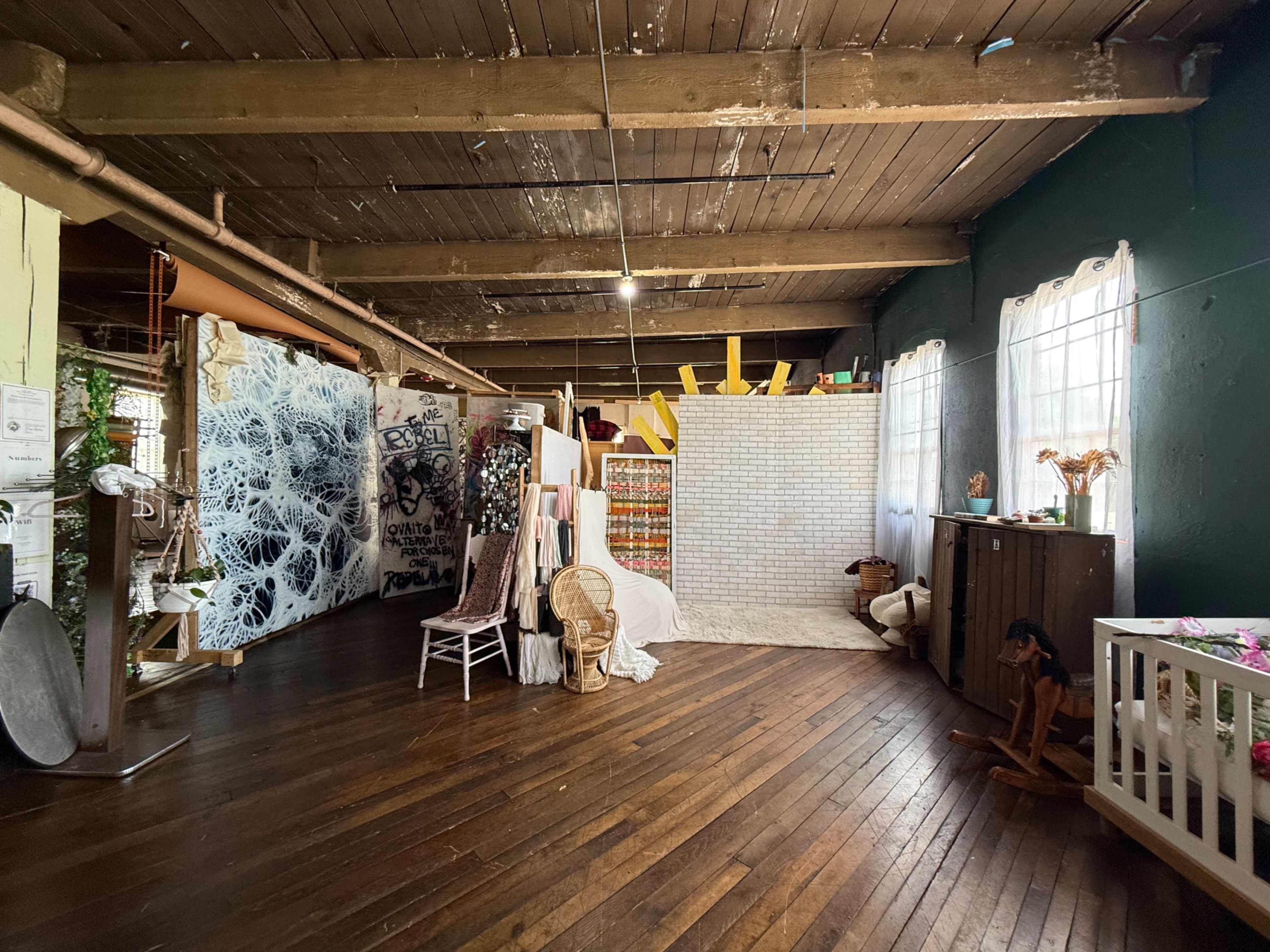 The image shows a spacious studio with wooden floors, featuring various decorative backgrounds, a wicker chair, and a corner with a crib.