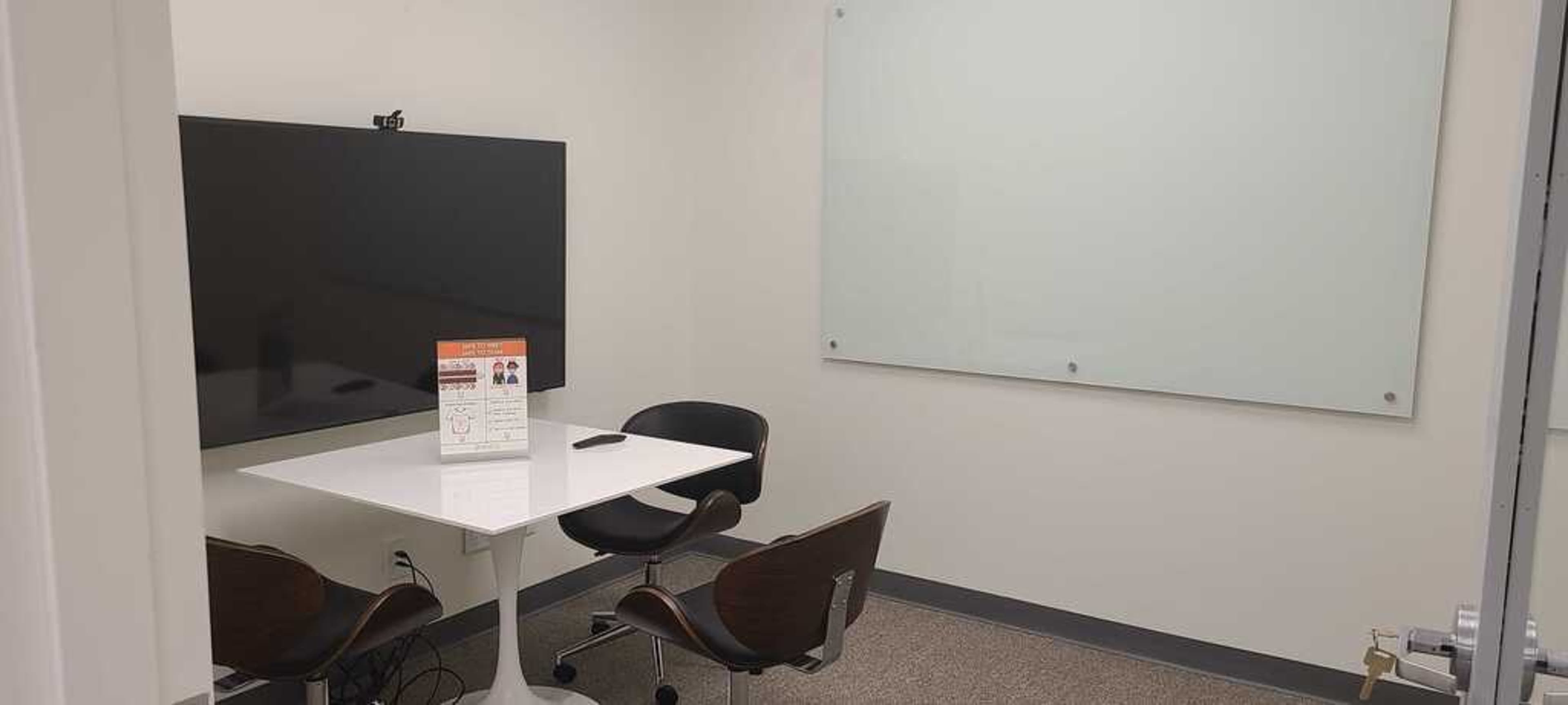 A small meeting room featuring a large television screen, a whiteboard, a table, and two chairs.
