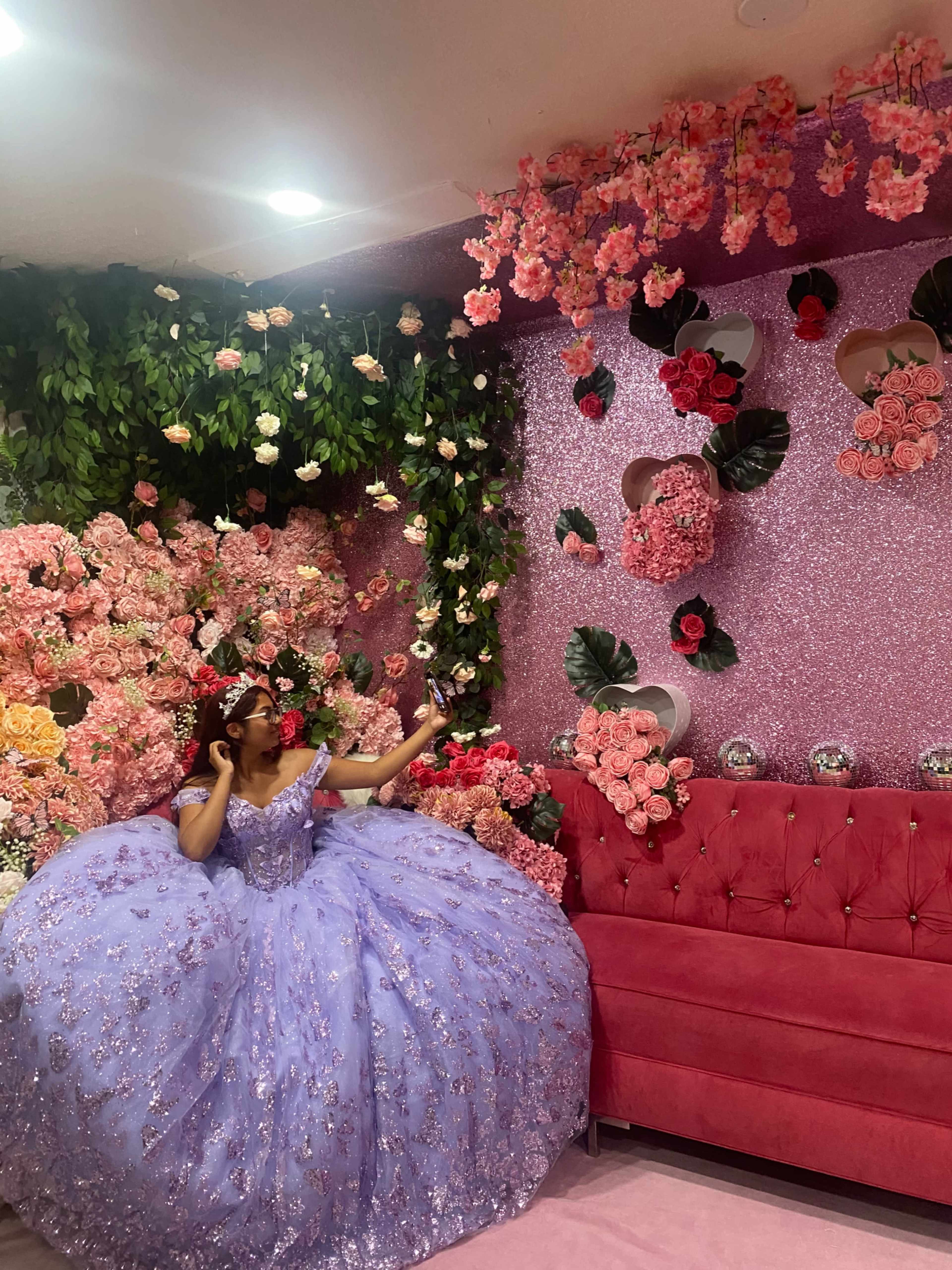 A person in a large purple gown sits on a pink tufted couch in a room adorned with floral decorations.
