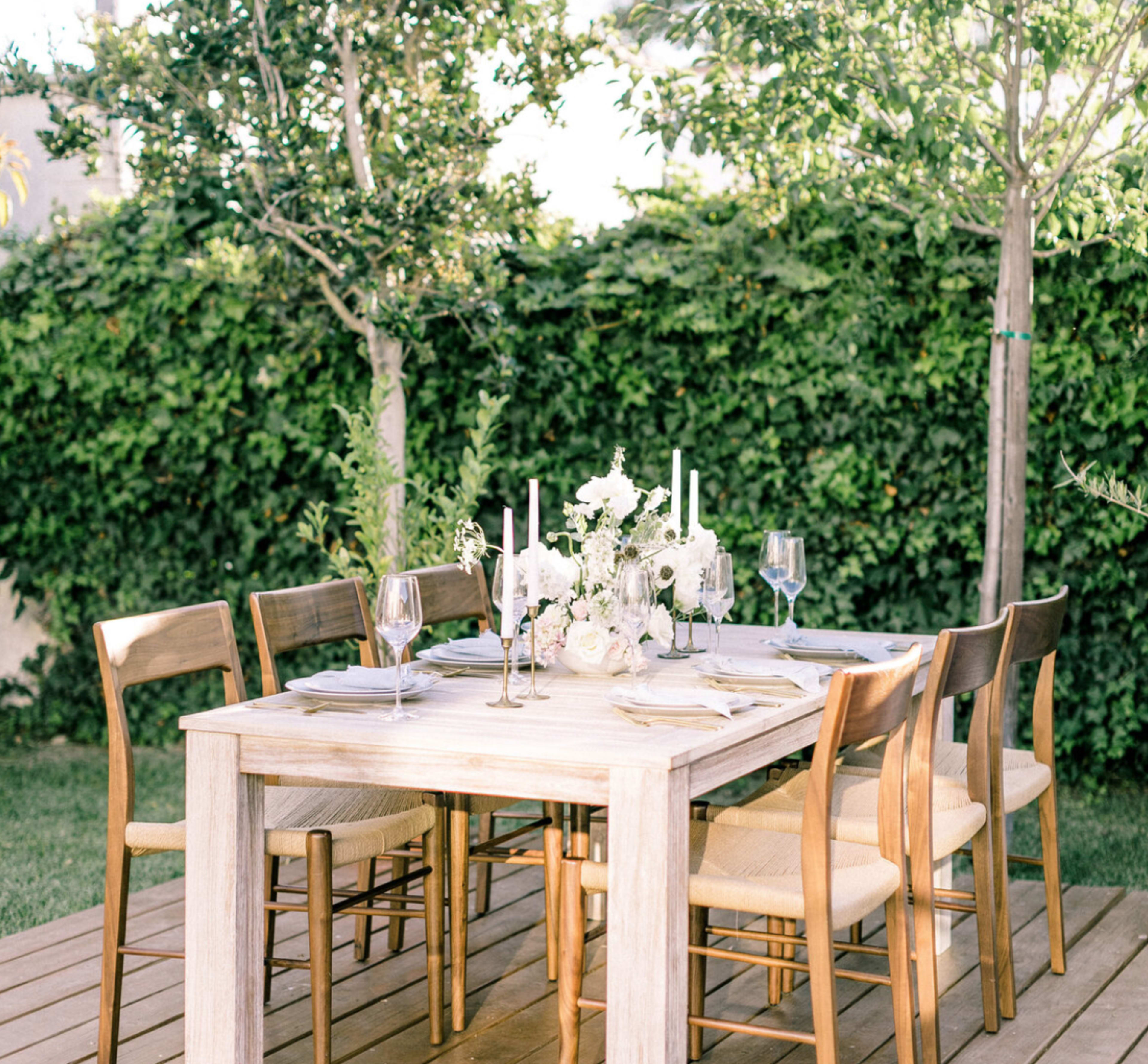 A wooden dining table is set outdoors, surrounded by chairs, with a floral centerpiece and candle holders atop a white tablecloth.