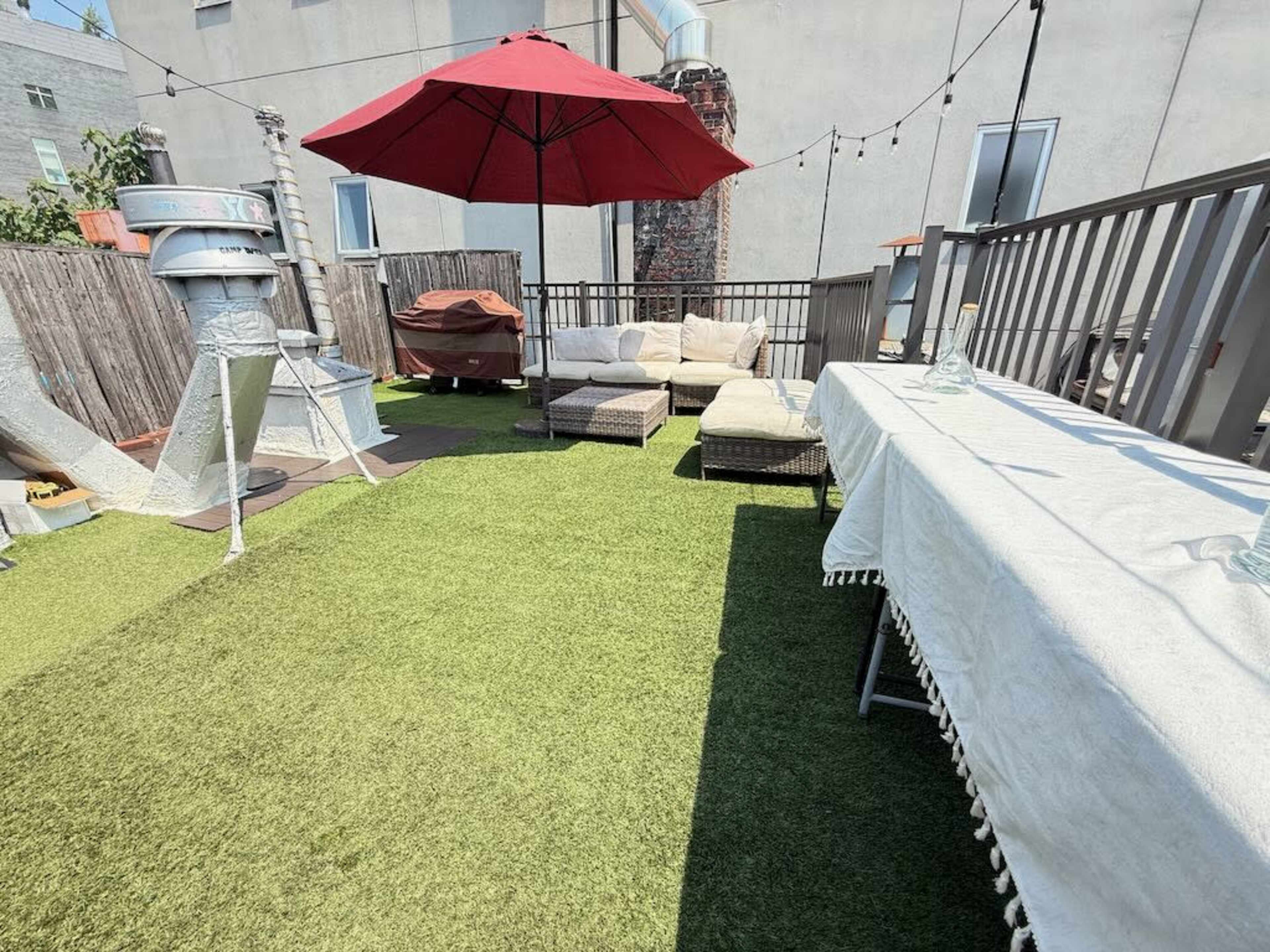 The image shows a rooftop terrace featuring a red umbrella over a seating area with lounge chairs and a table, surrounded by green artificial grass.