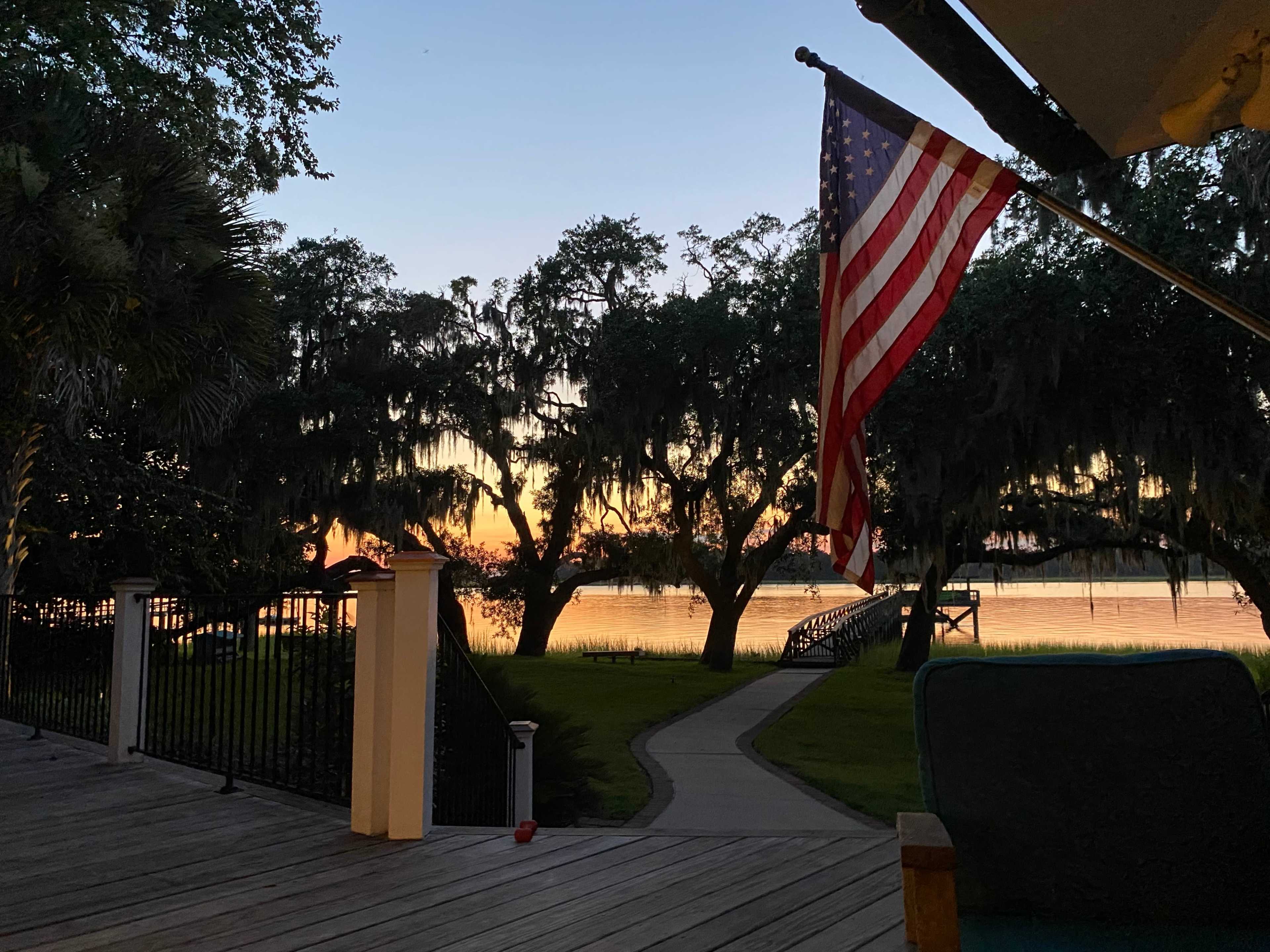 The image shows a wooden deck with an American flag waving gently, overlooking a lake at sunset with trees and a winding path in the background.