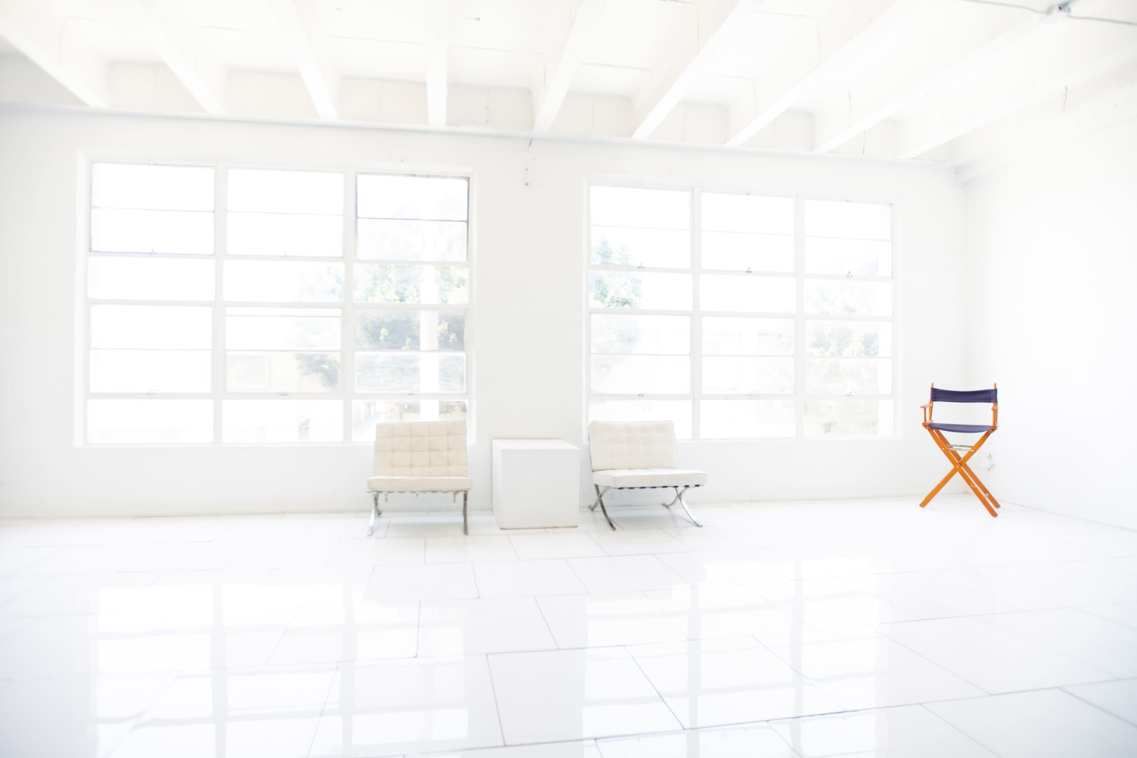 The image shows a bright, minimalist interior with two white chairs, a small white table, and a single orange director's chair beside large windows.
