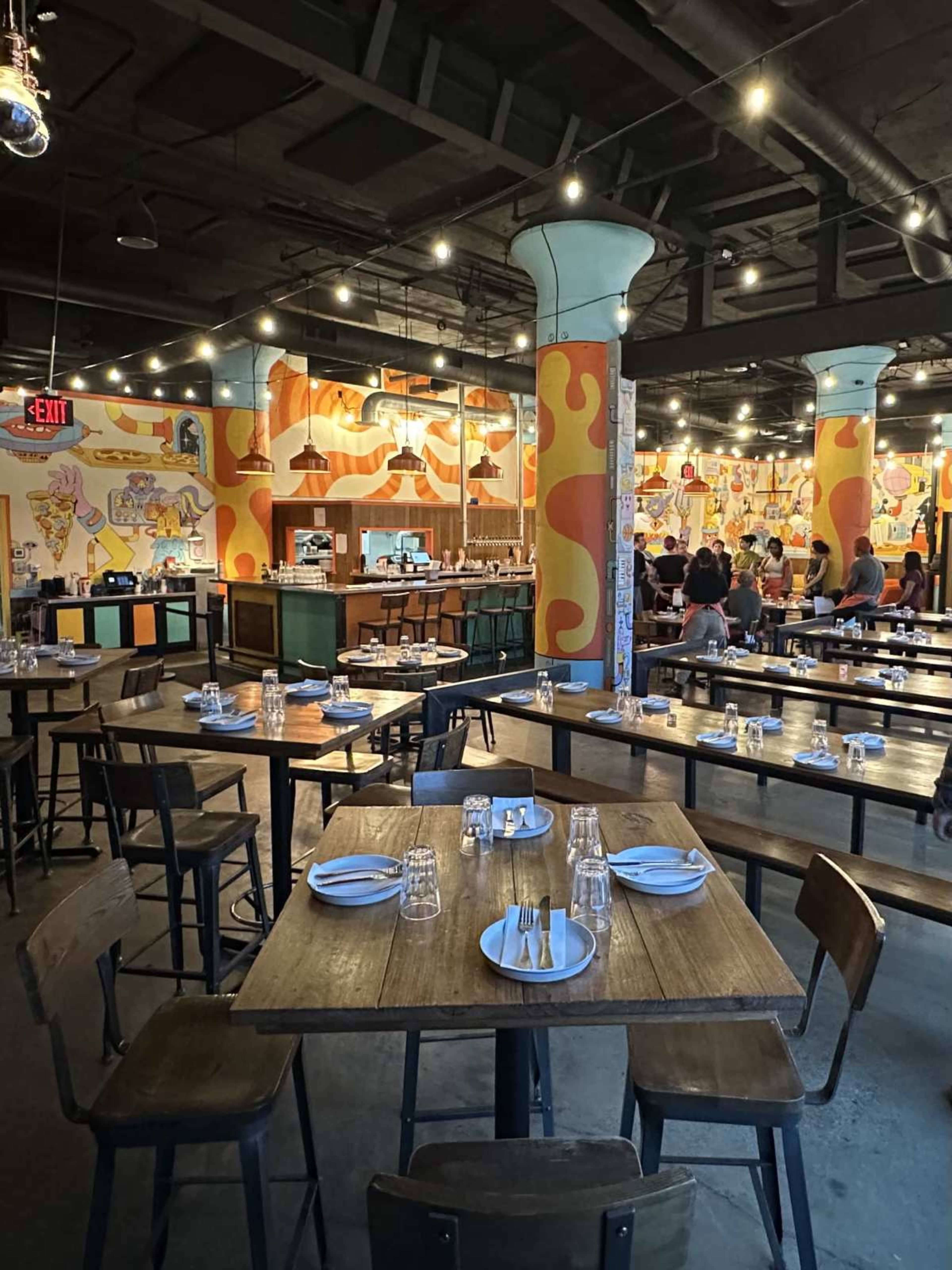 The image shows a vibrant restaurant interior with colorful murals on the walls, wooden tables set for dining, and overhead string lights illuminating the space.