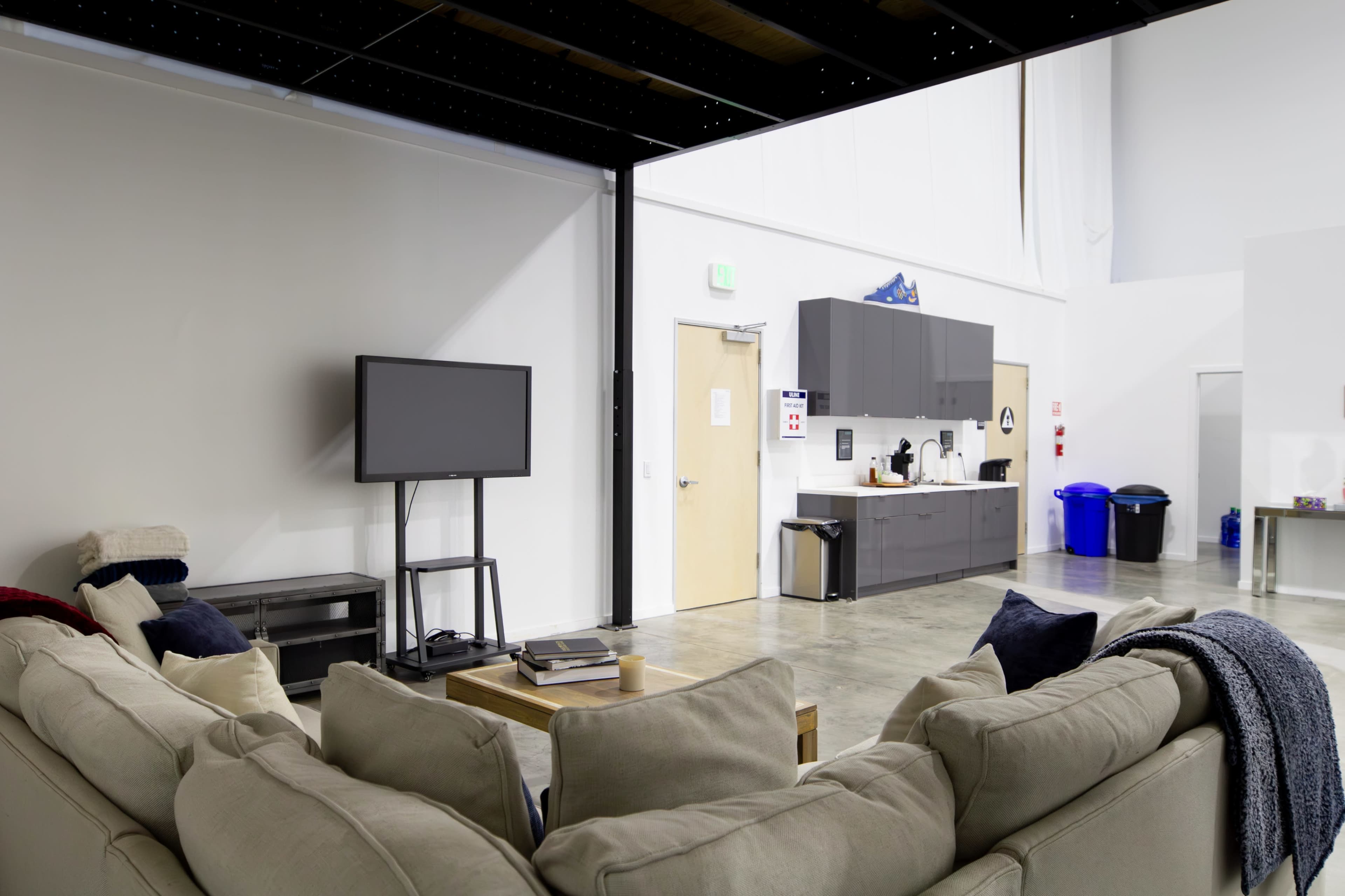 The image shows a spacious, modern shared living area with a couch, a television, and a kitchen space featuring cabinets and appliances.