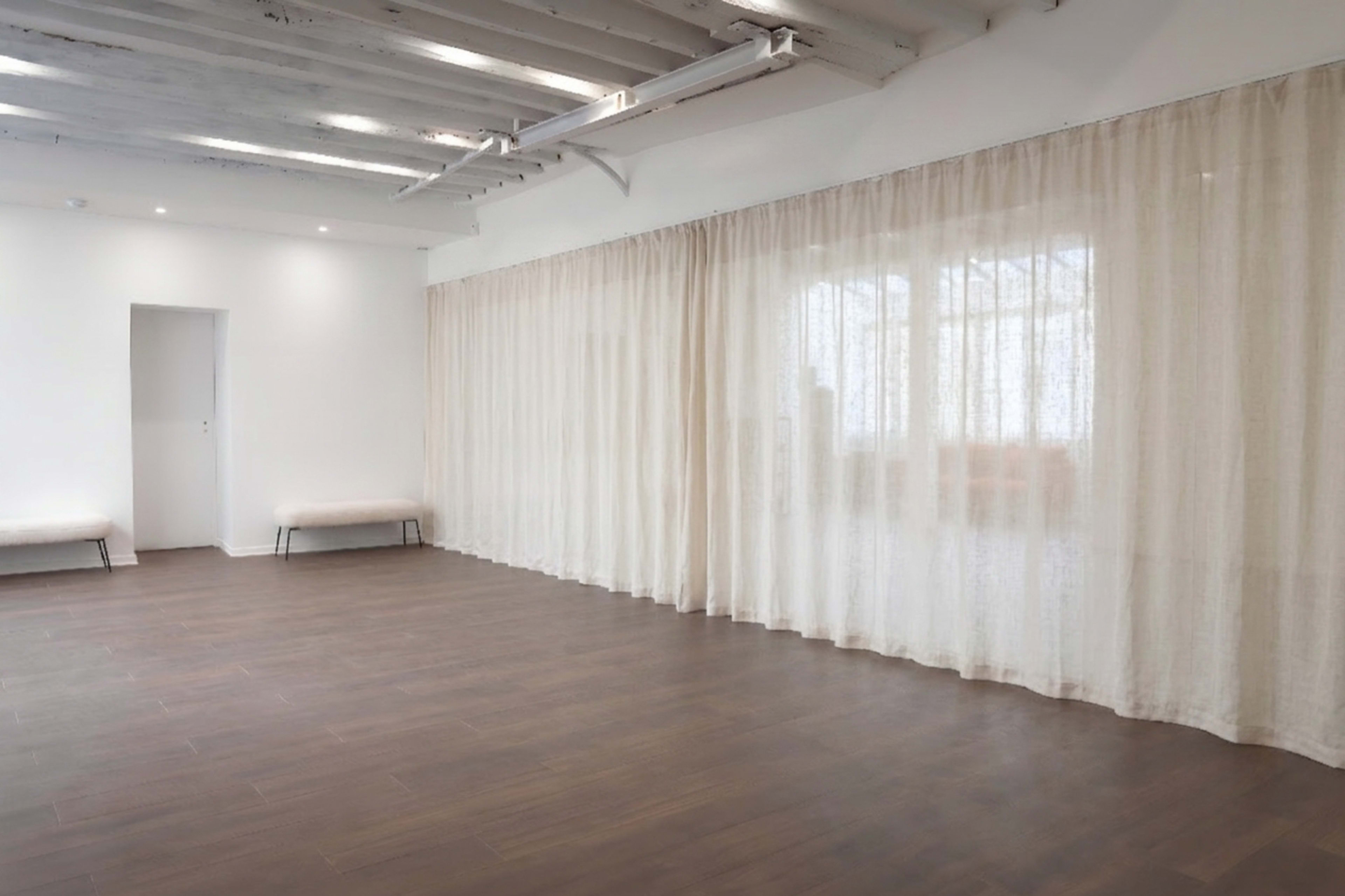The image shows an empty, well-lit room with wooden flooring and sheer curtains along one wall.