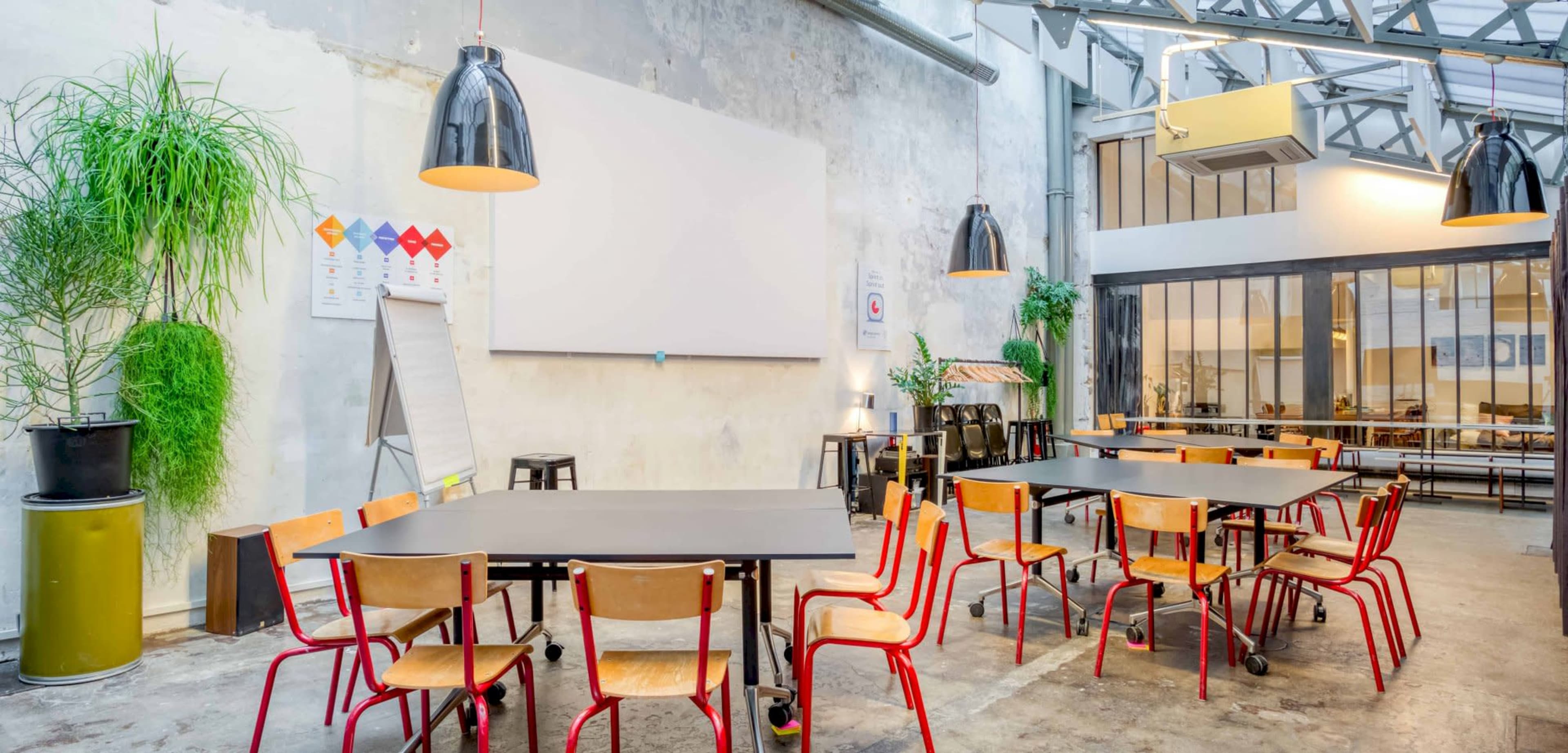 A well-lit meeting room features a large screen, black tables, and red-legged chairs arranged for a group setting.