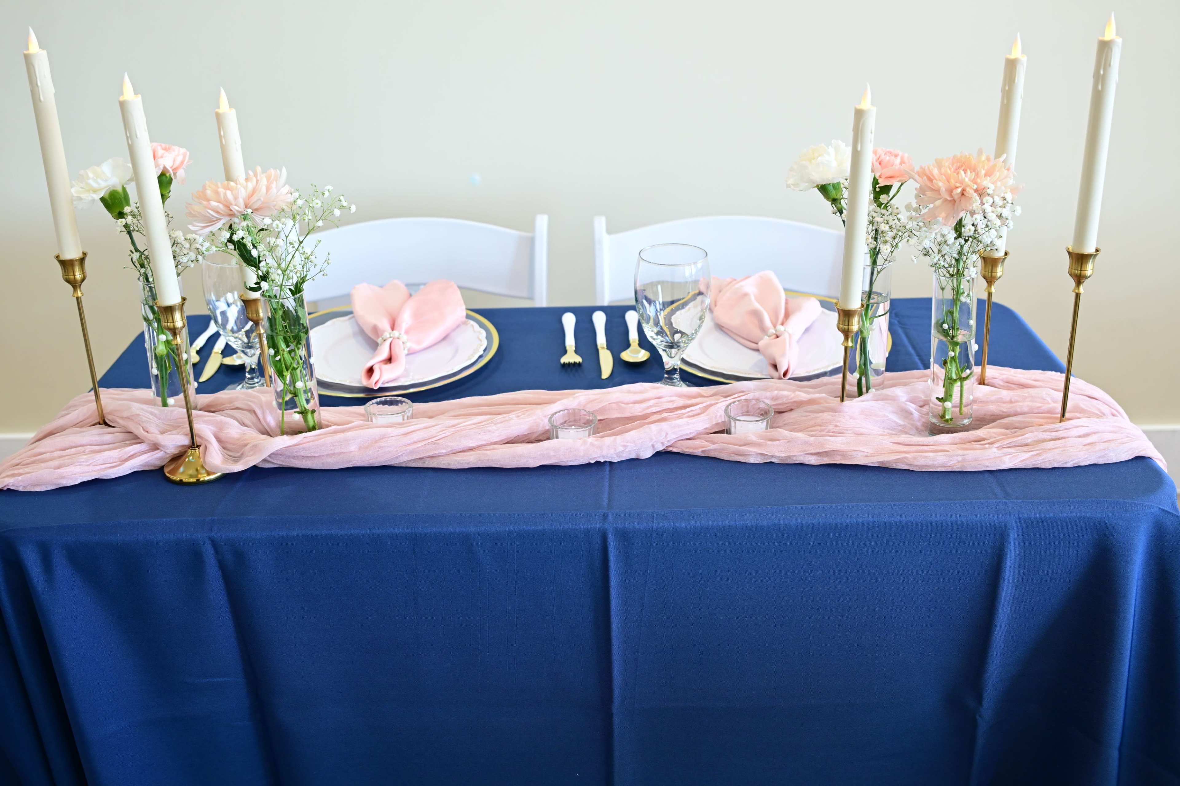 A dining table is elegantly set with a navy blue tablecloth, plates, glasses, and floral arrangements, adorned with pink napkins and candle holders.
