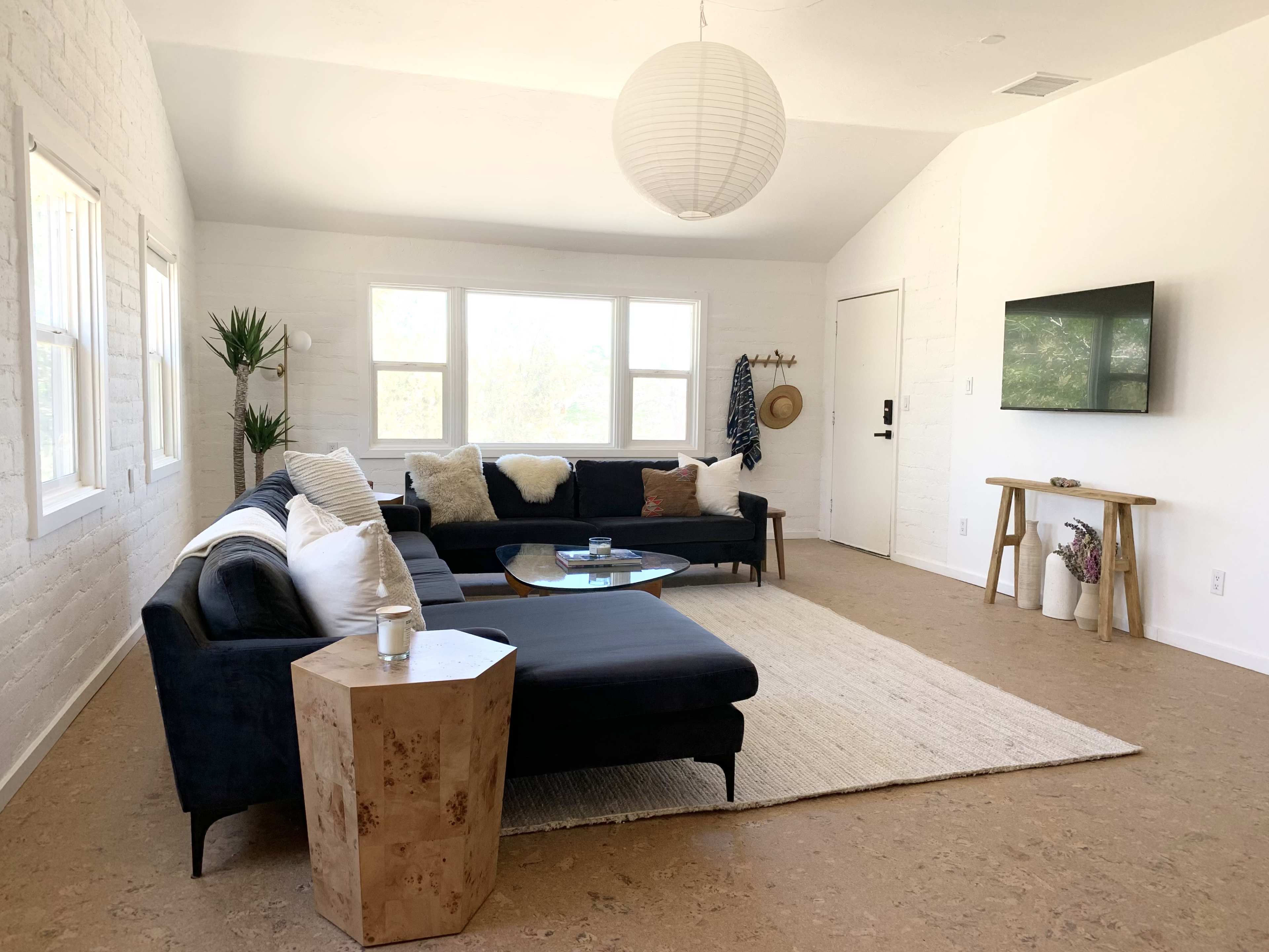 A modern living room featuring a black sectional sofa, a glass coffee table, a light-colored rug, and large windows allowing natural light to fill the space.