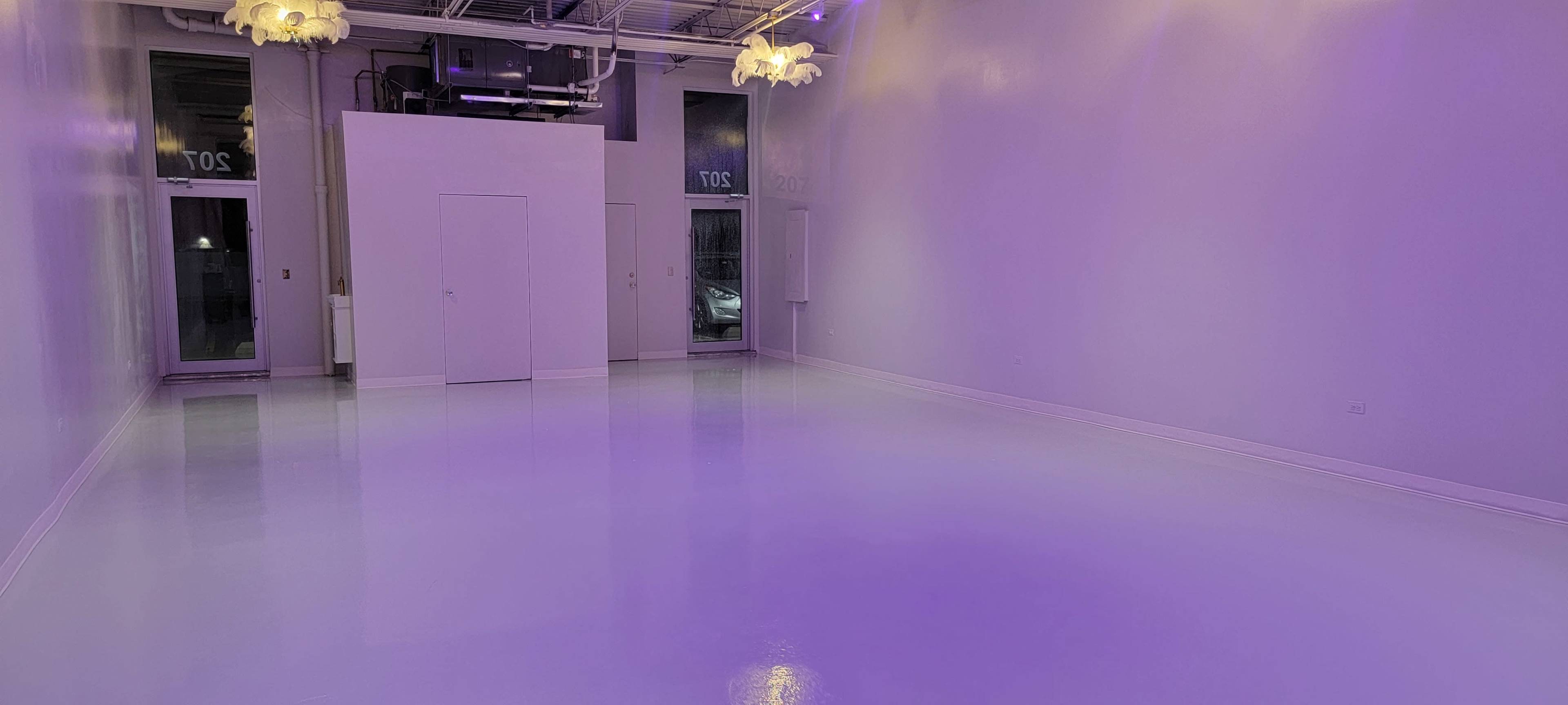 The image shows a large, minimally furnished room with glossy white floors and purple lighting, illuminated by hanging light fixtures.