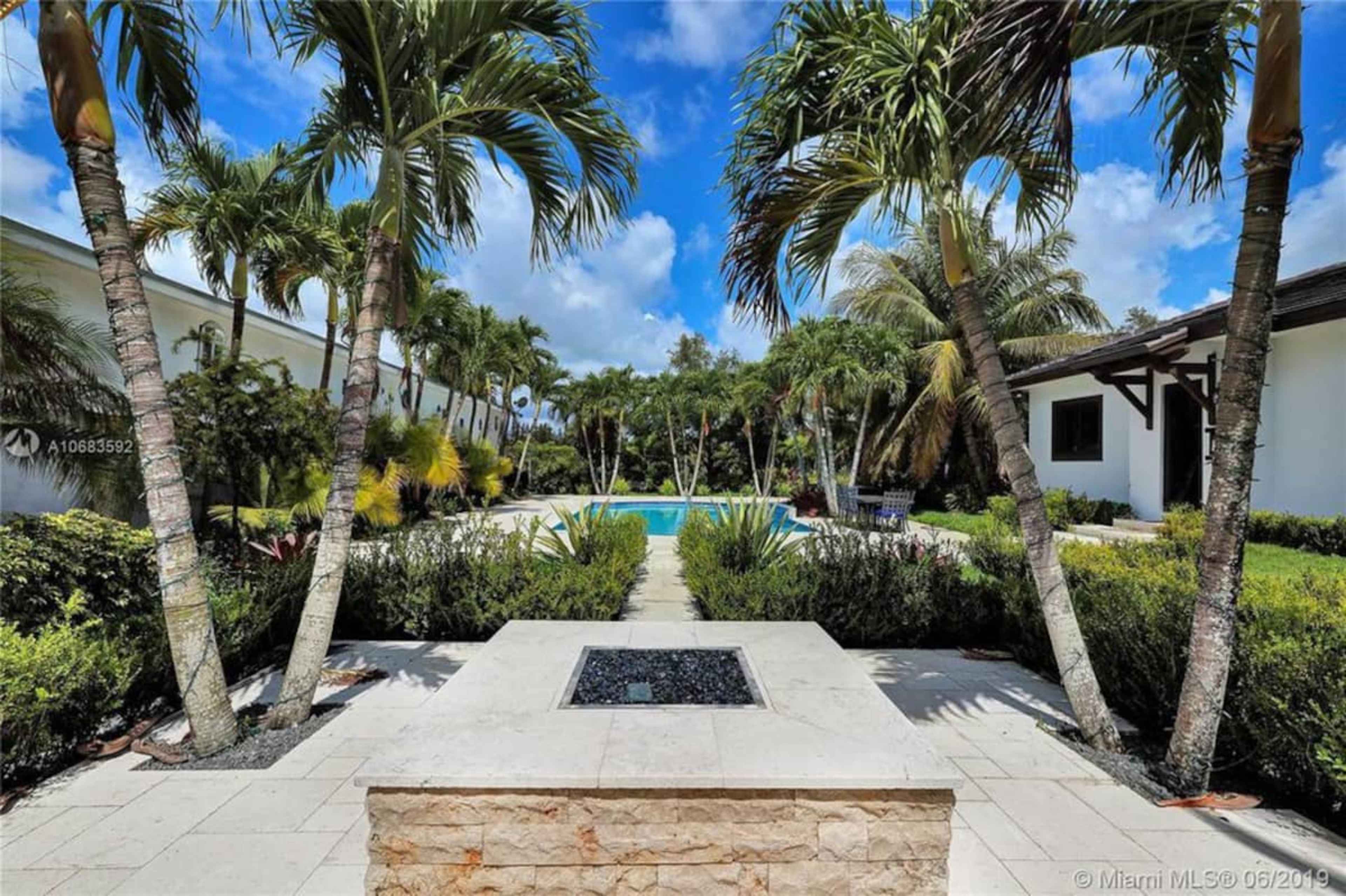 A view of a landscaped backyard featuring a pool, palm trees, and a stone fire pit.