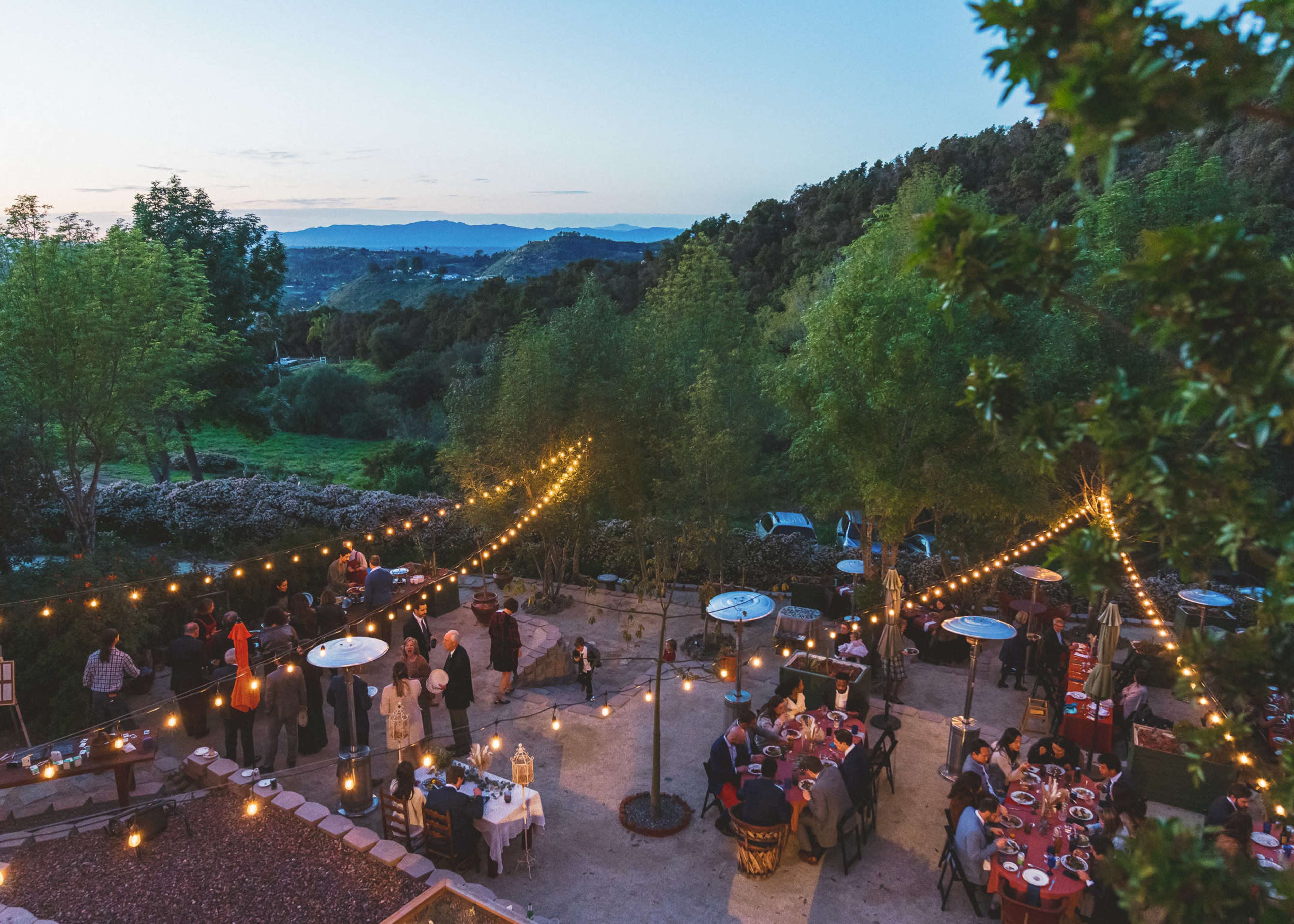 A large outdoor gathering is set in a scenic location with string lights illuminating tables and guests enjoying food and drinks as the sun sets over the hills.