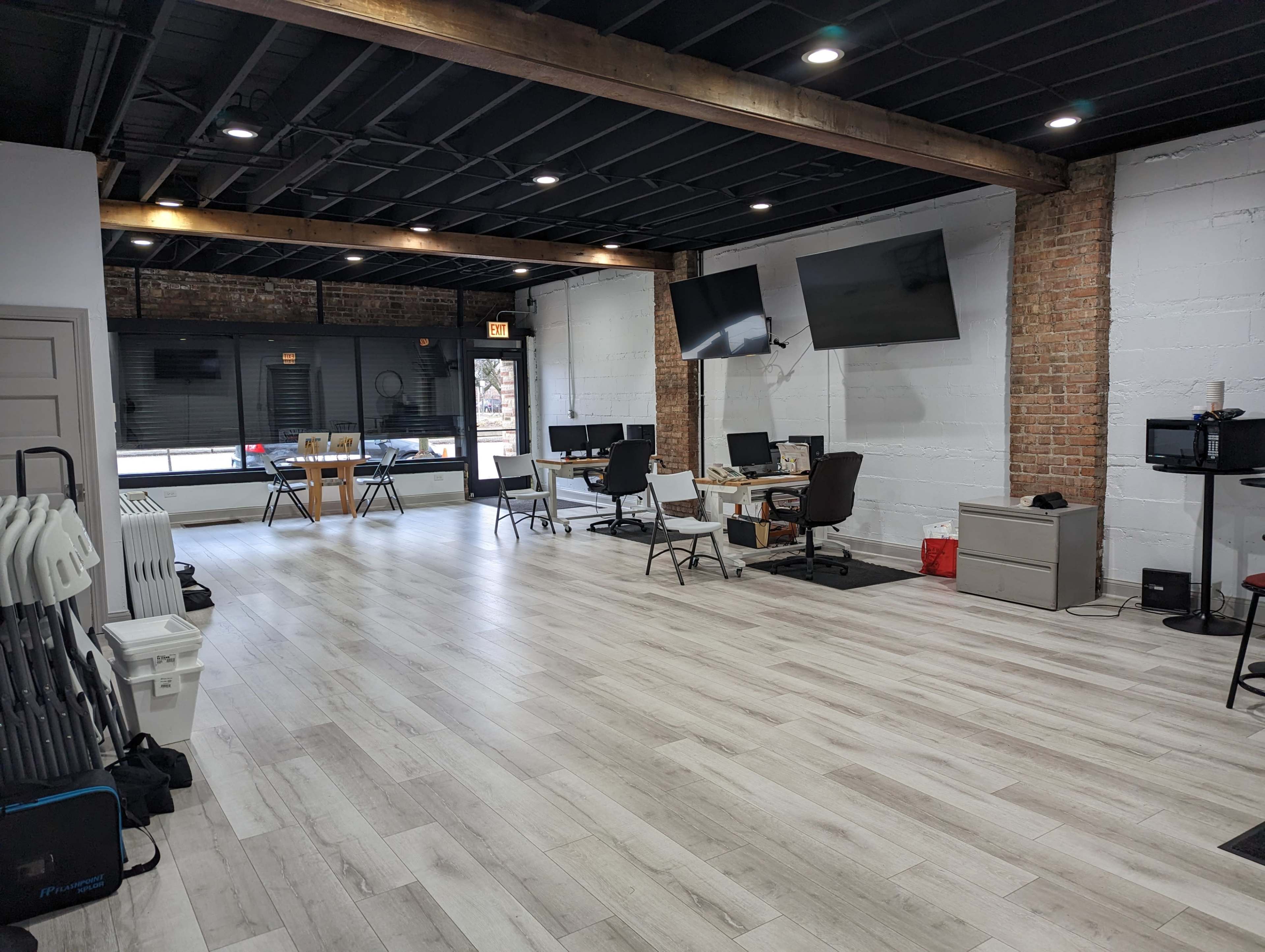 A spacious office environment with several desks, chairs, and large windows, featuring exposed brick walls and wooden beams.