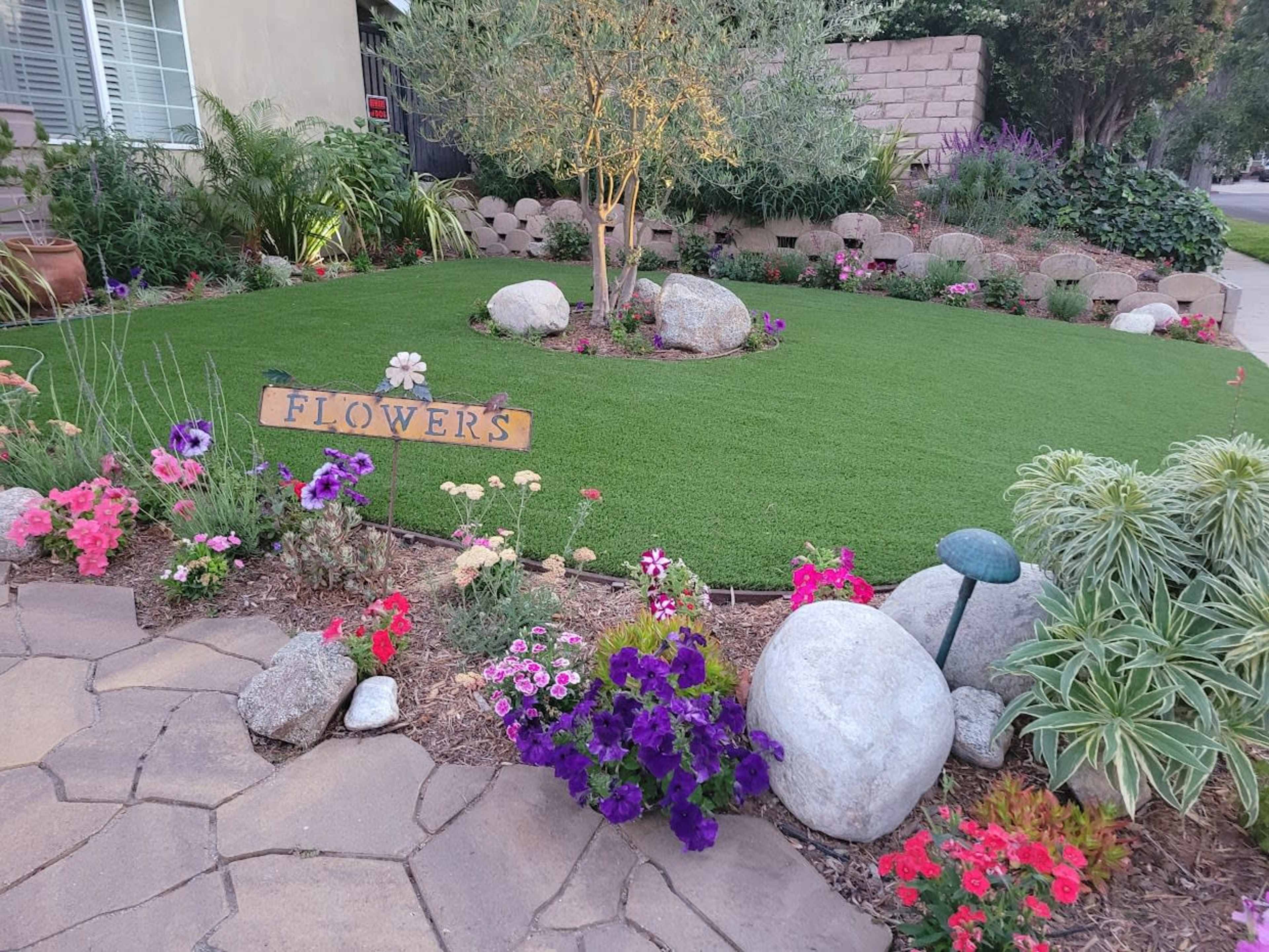 The scene features a landscaped front yard with artificial grass, colorful flower beds, decorative rocks, and a wooden sign that reads "Flower's."