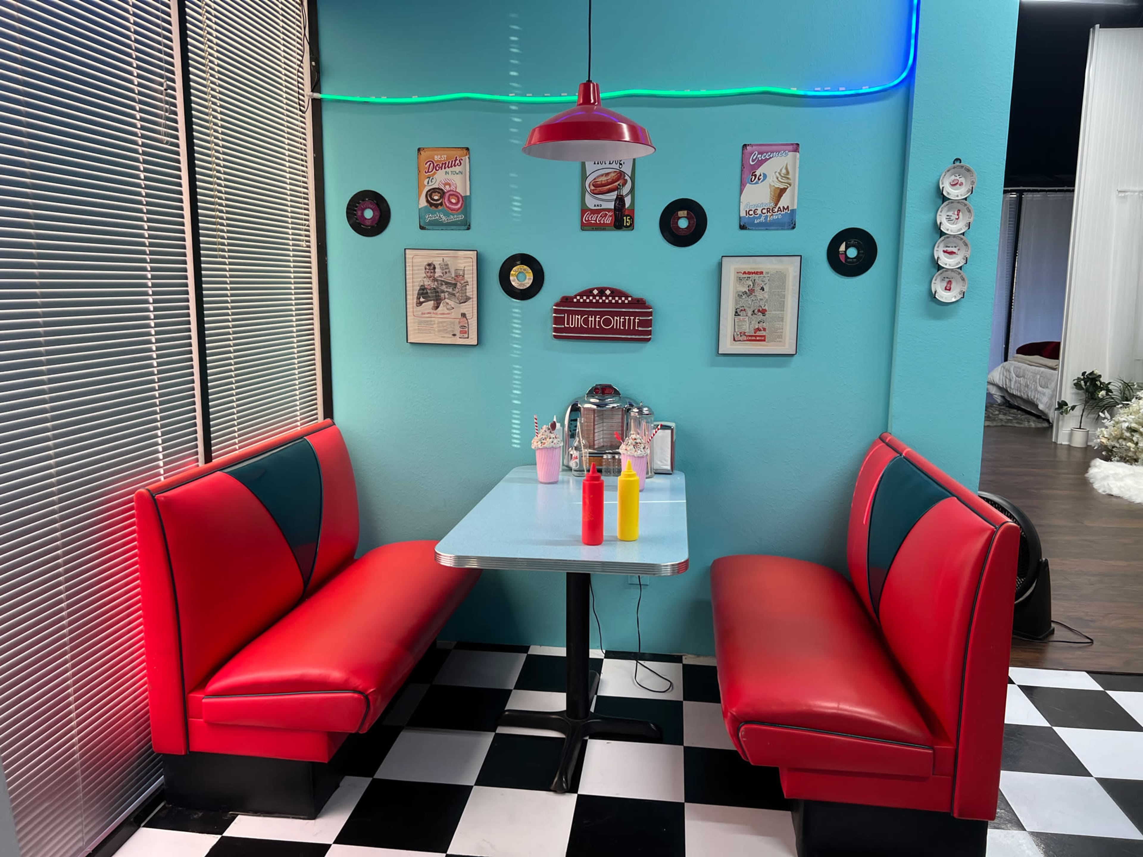 The image shows a retro-style diner booth with red vinyl seating and a checkered floor, featuring a small table set with condiments and decorated with vintage records and posters on the walls.
