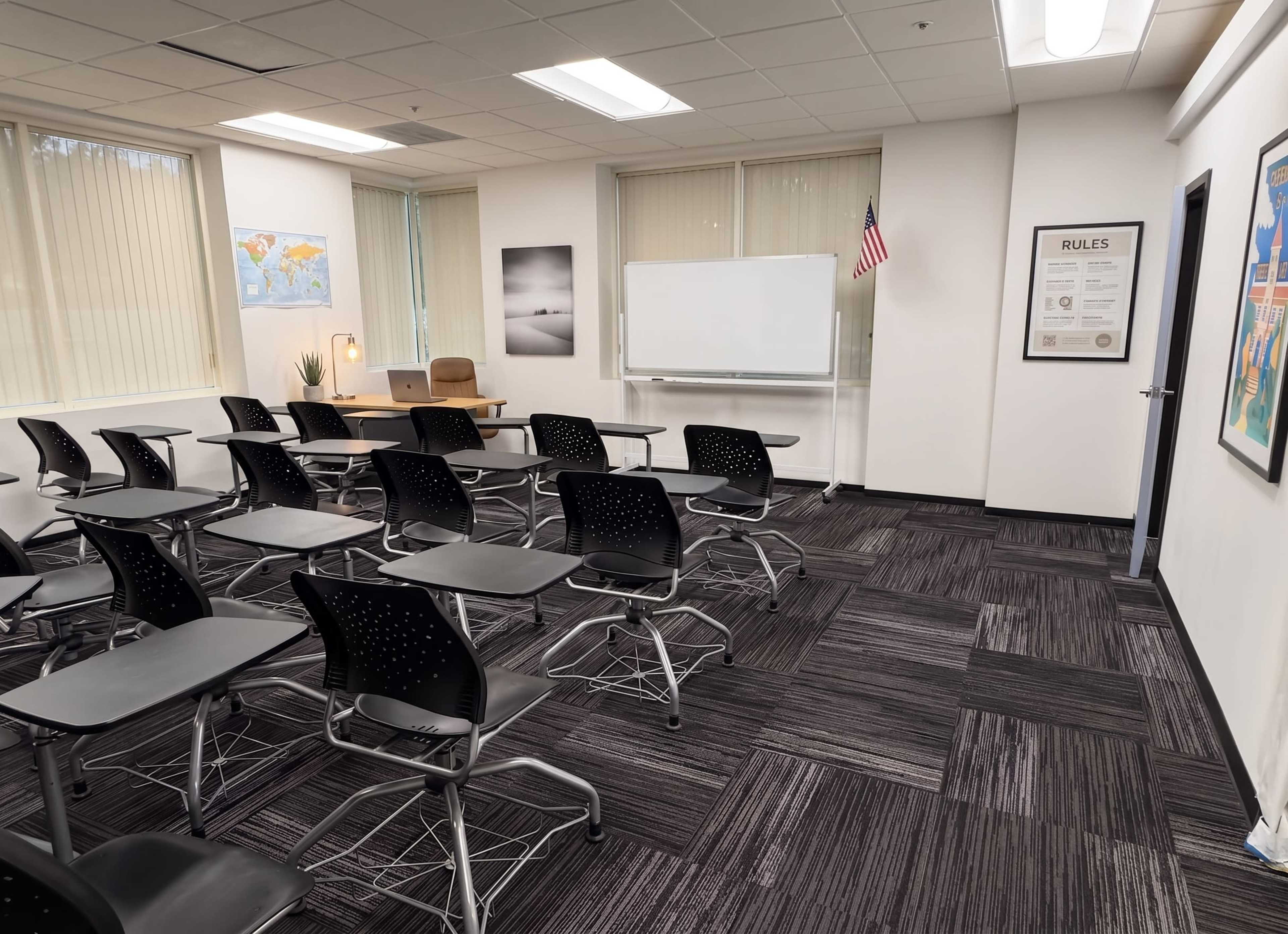 The image shows a classroom with rows of black chairs facing a large whiteboard, a desk with a chair, and educational posters on the walls.