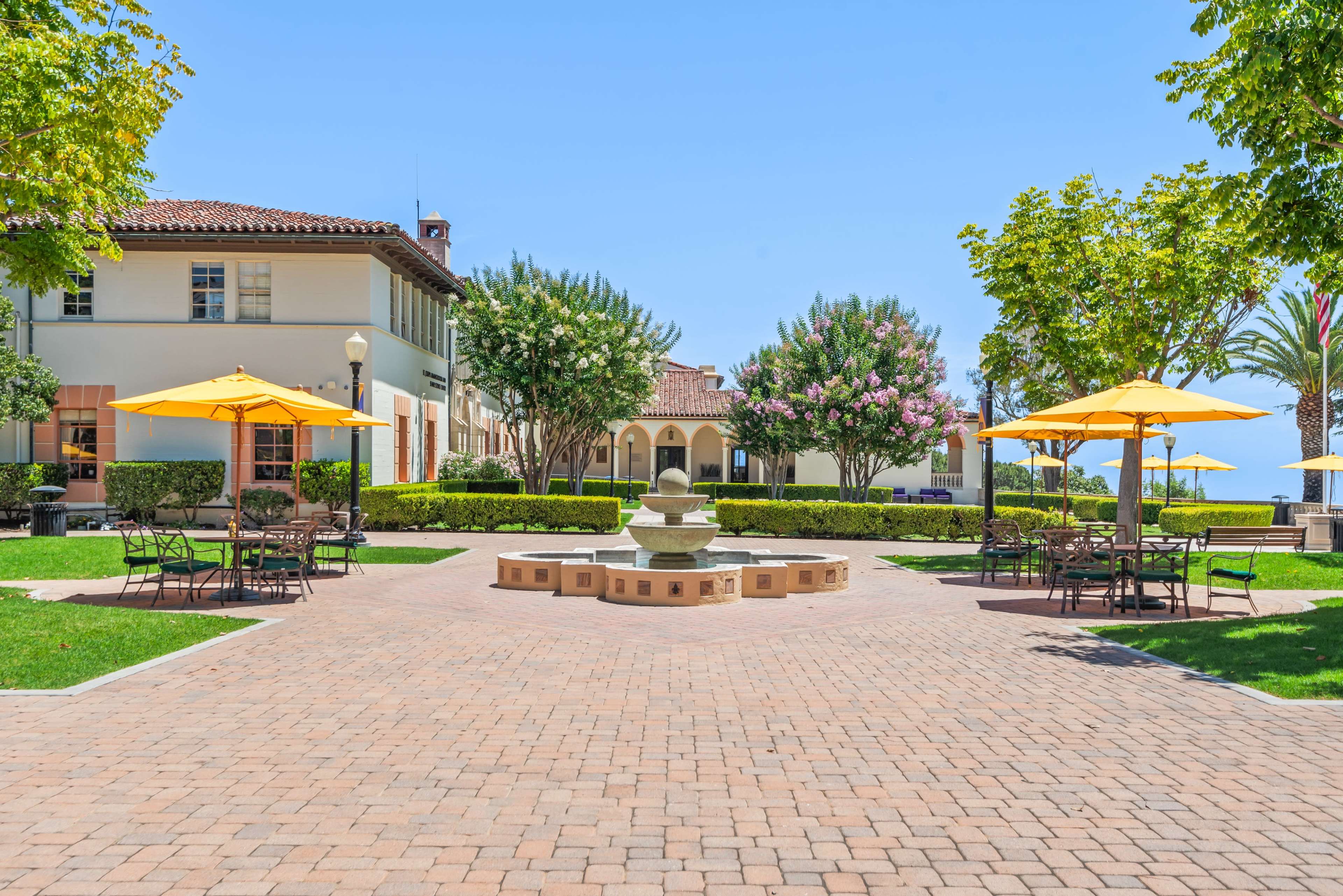 The scene features a courtyard with a central fountain surrounded by neatly arranged tables and umbrellas, flanked by well-maintained greenery and buildings in the background.