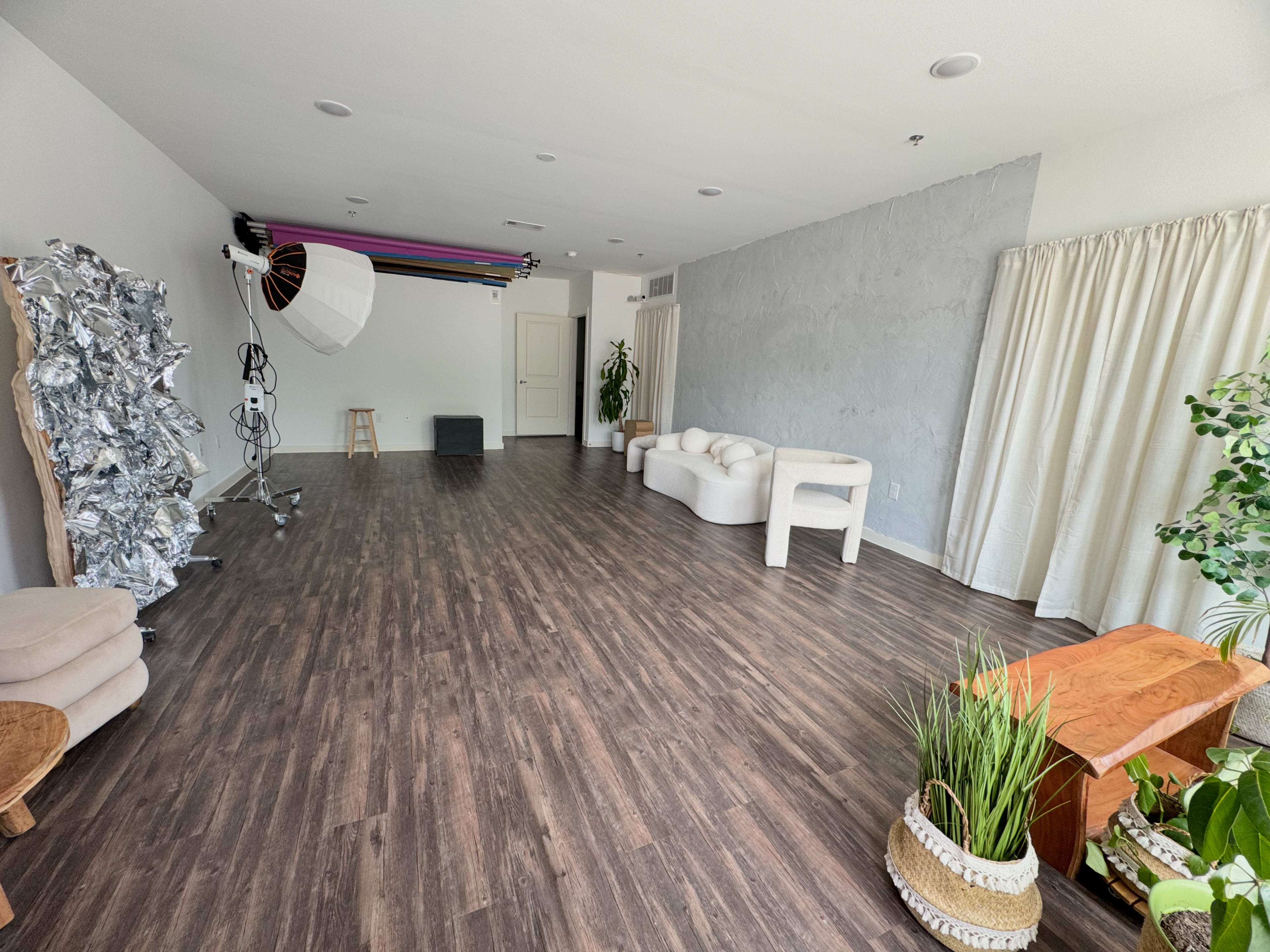 The image shows a spacious, brightly lit studio with wooden flooring, minimalistic furniture, and photography equipment in one corner.