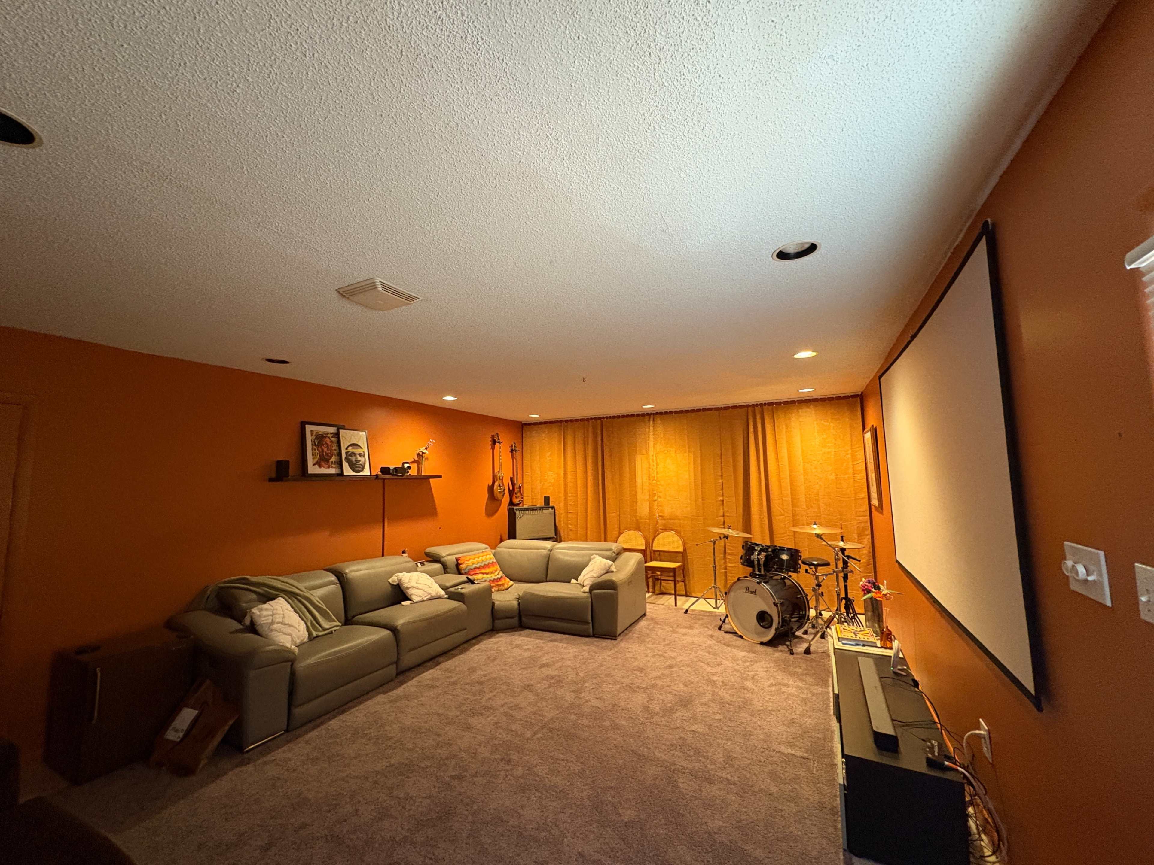 The image shows a cozy basement living area with a sectional couch, a large screen, a drum set, and warm orange walls.
