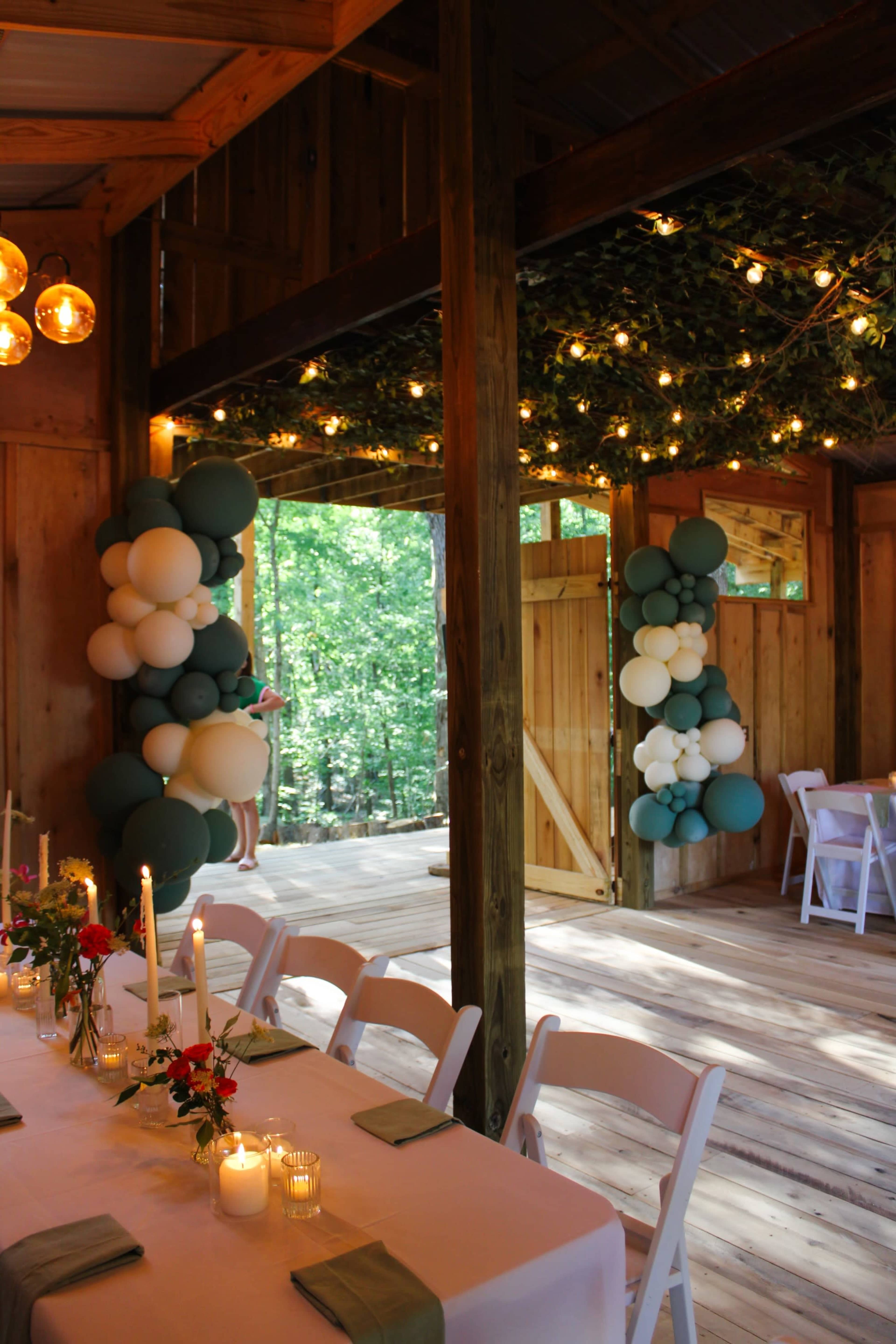 The interior of a wooden venue is decorated with balloons and string lights, featuring tables set with flowers and candles.