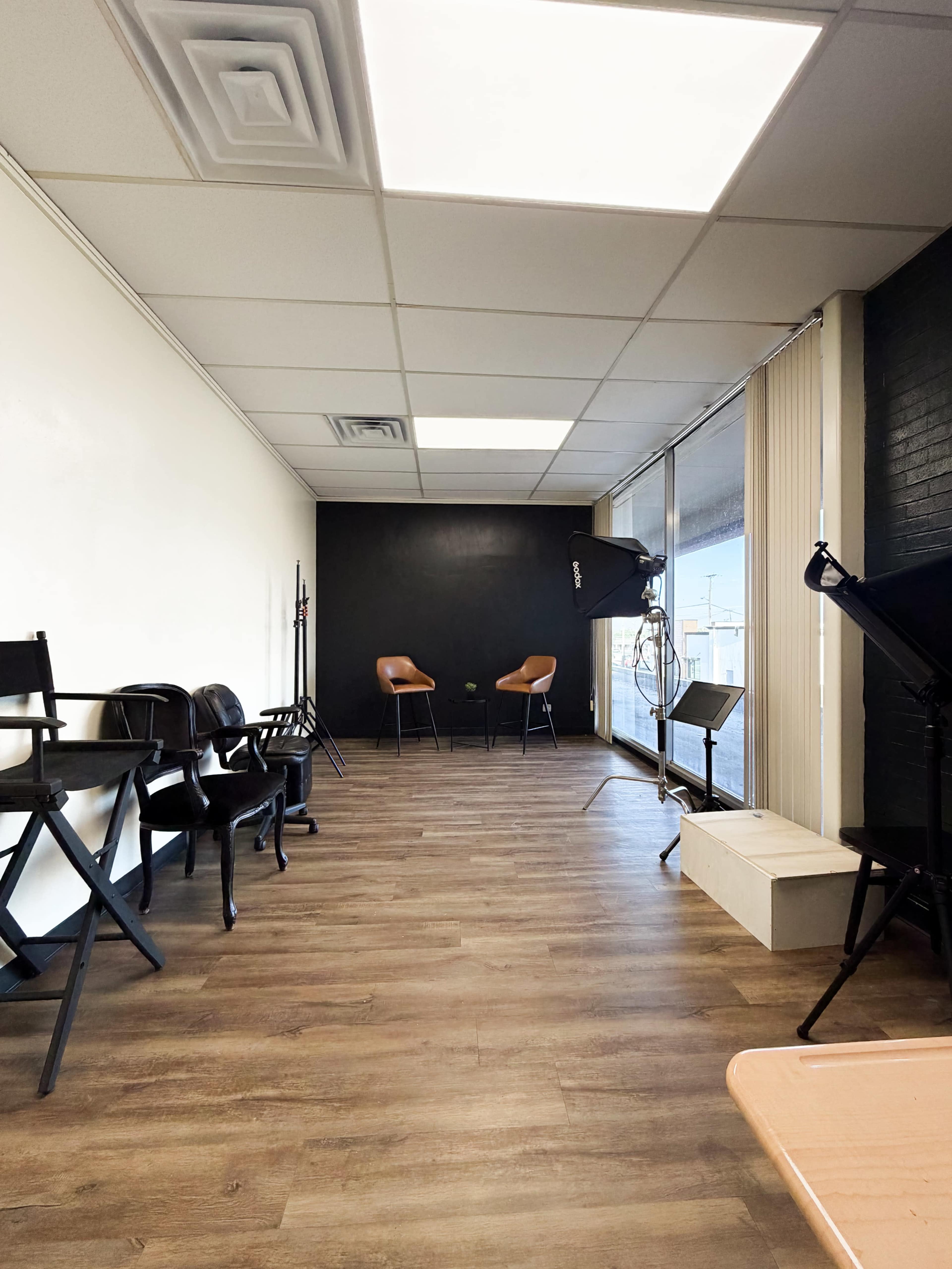 The image shows a spacious room with wooden flooring, featuring two brown chairs, black walls, and several foldable chairs lined up against one side.