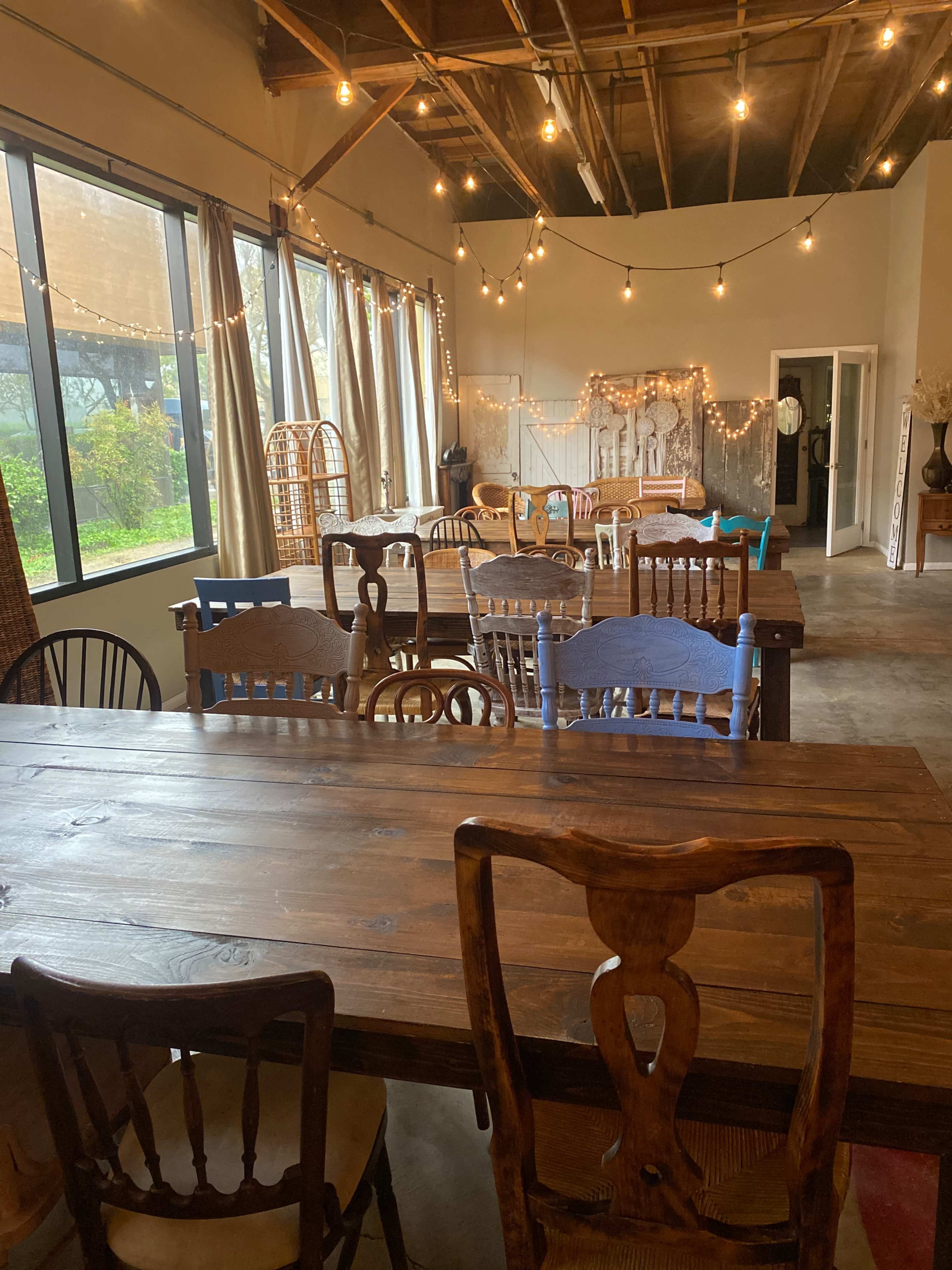 The image shows a spacious interior room with wooden tables and chairs, large windows, and string lights overhead.
