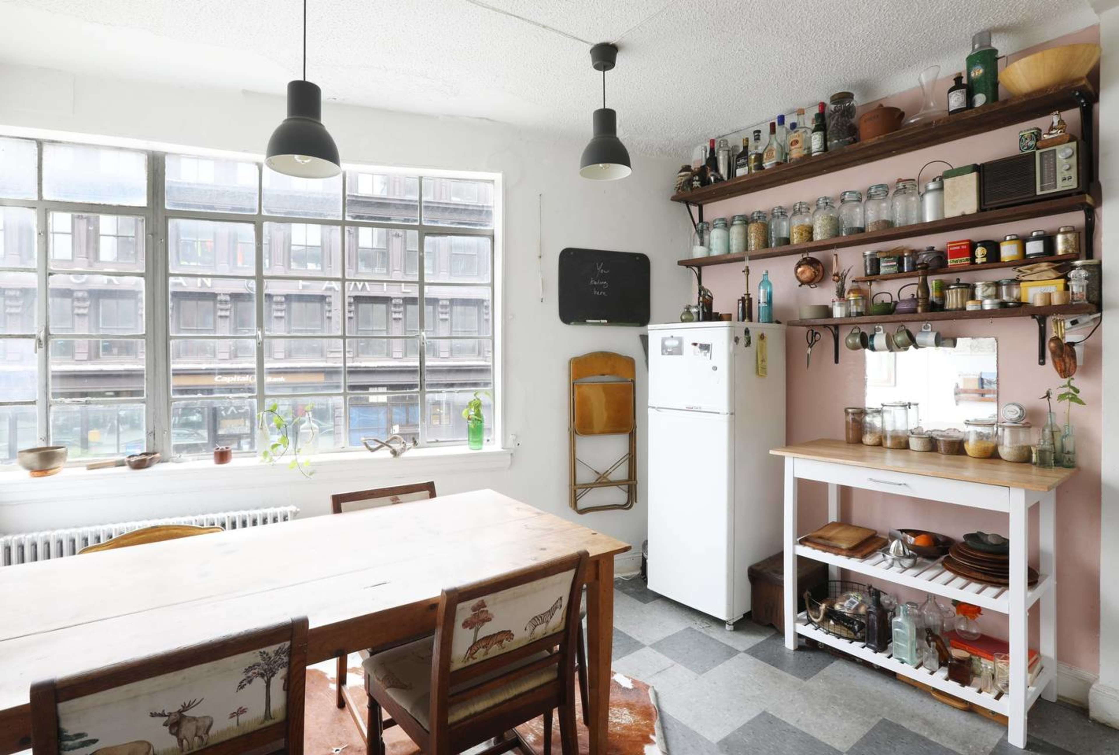 A bright kitchen features a wooden dining table, a white refrigerator, and shelves filled with jars and kitchenware, all under industrial-style pendant lights.
