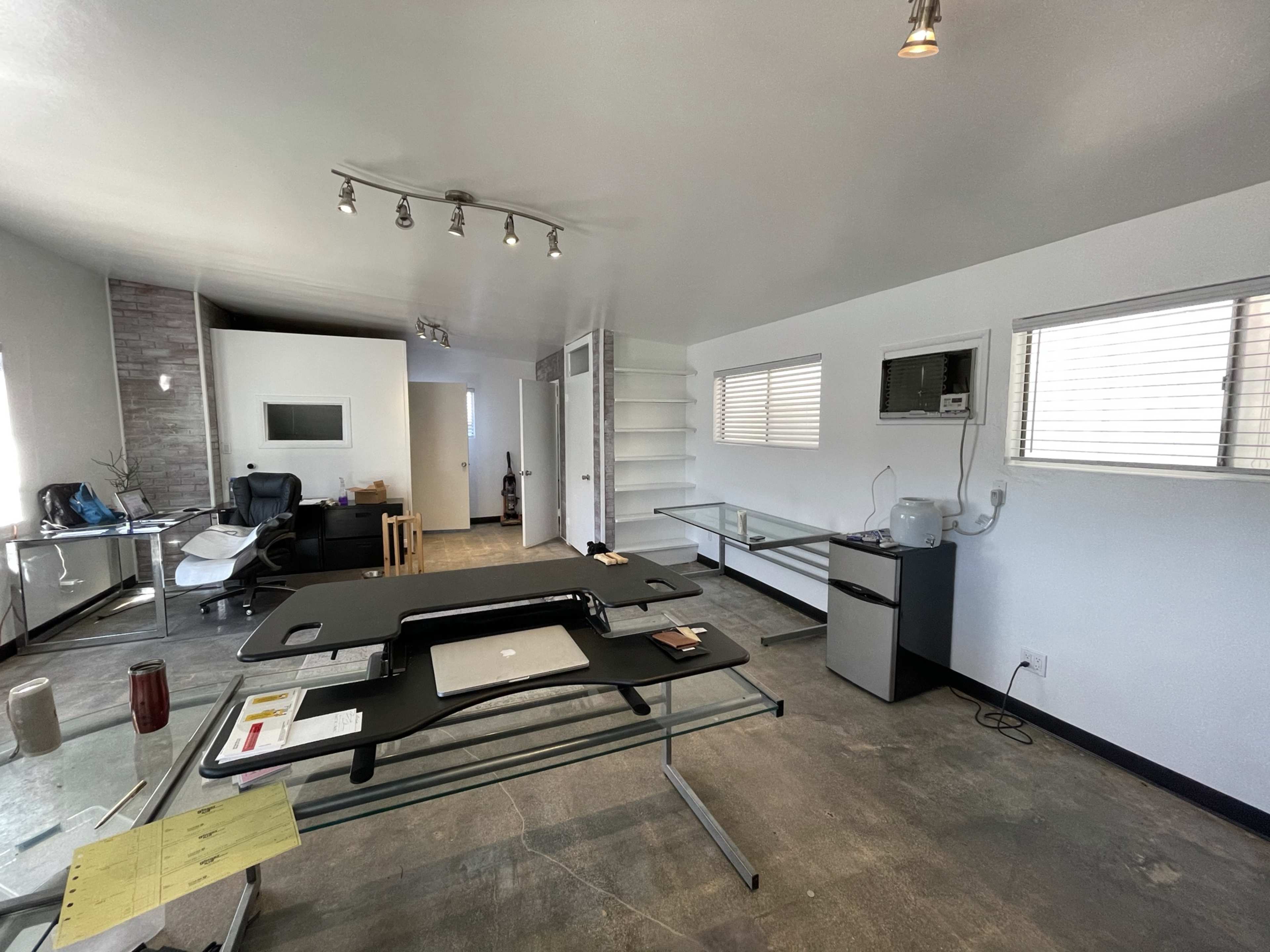 The image shows a modern, minimalistic office space with a glass desk, a chair, shelves, and an air conditioning unit.