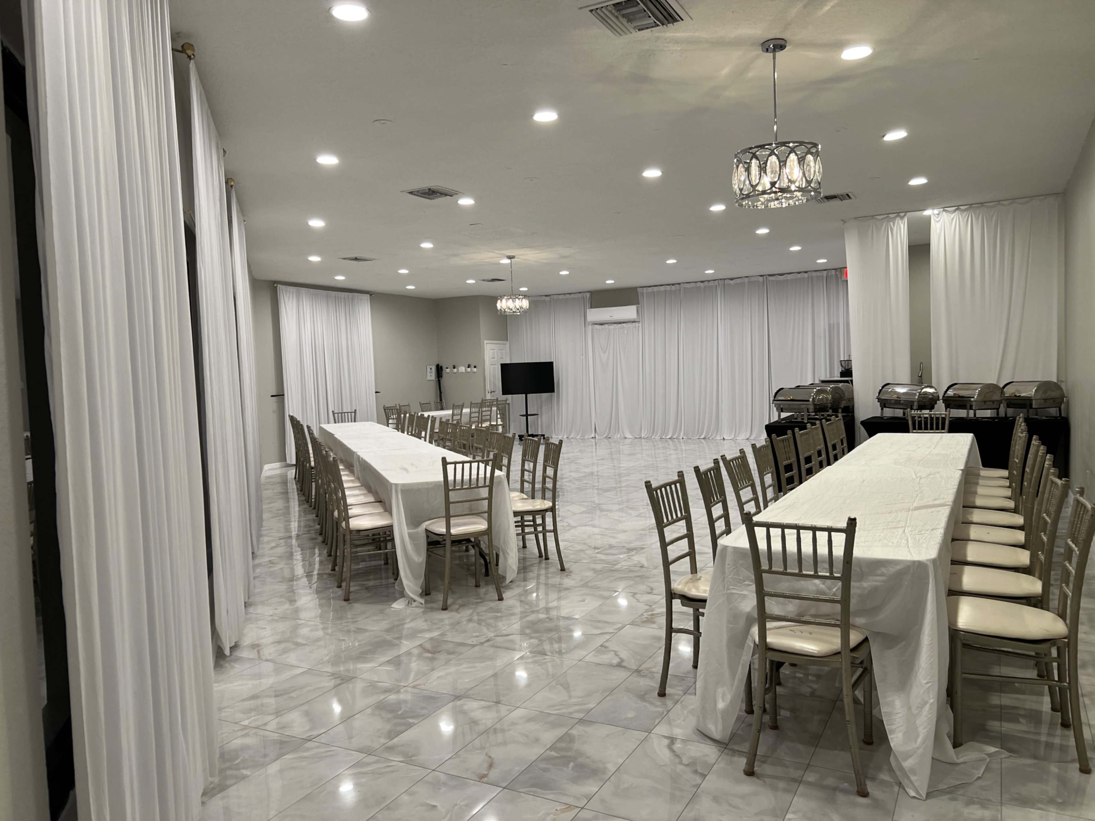 The image shows a spacious, elegantly arranged dining area with long tables covered in white cloth and gold chairs, surrounded by white drapes.