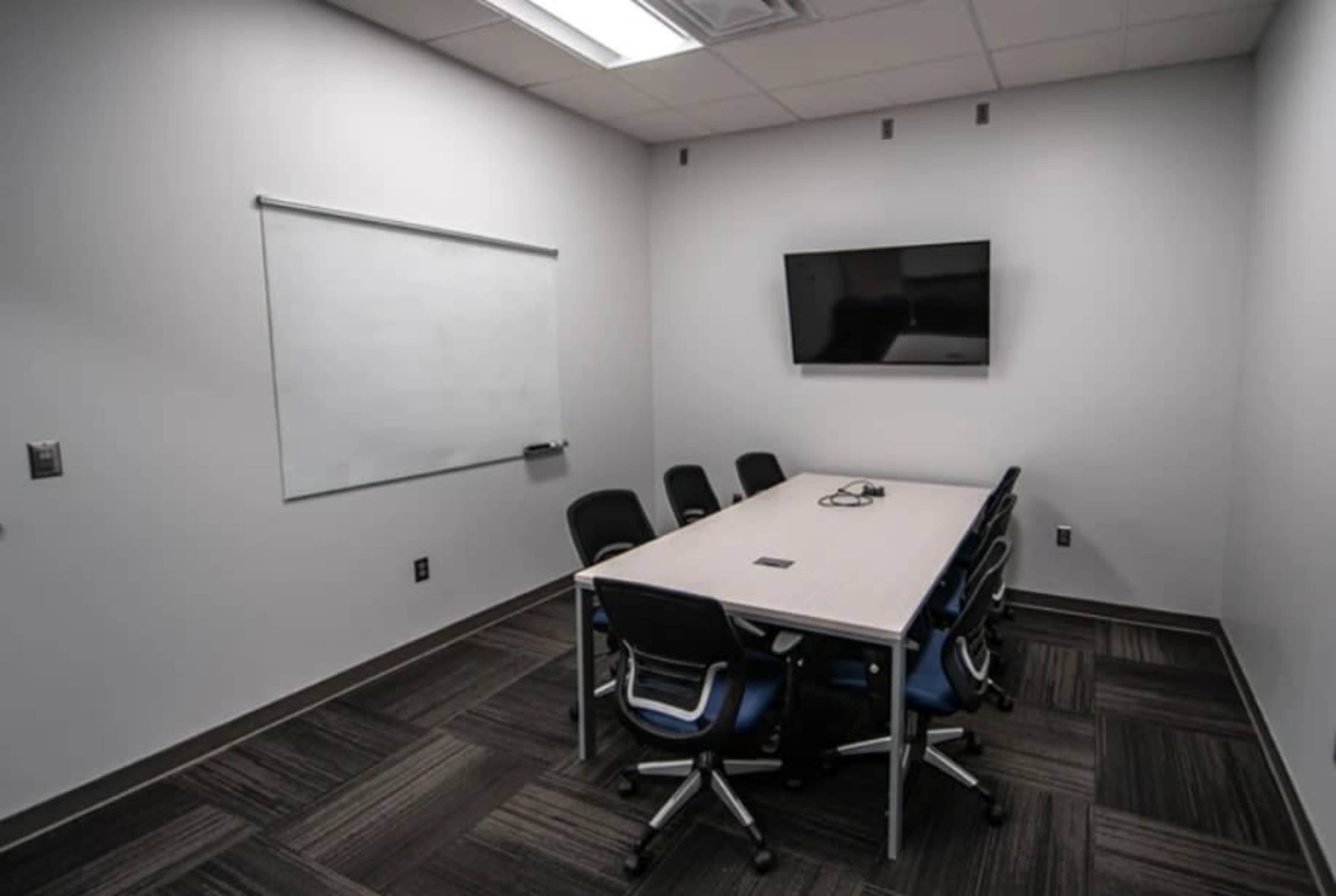 A small conference room features a whiteboard, a wall-mounted television, and a rectangular table surrounded by six chairs.