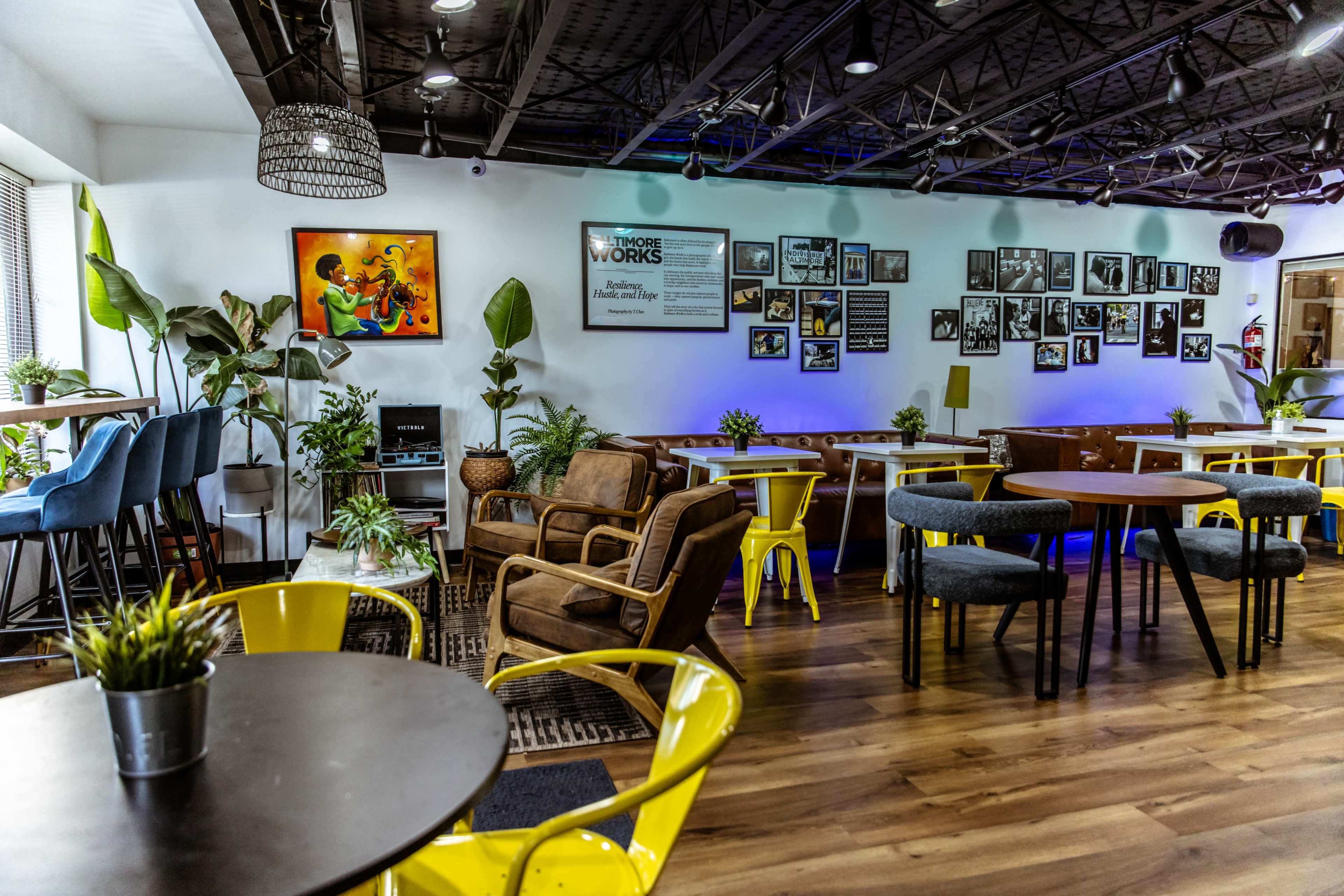 The image shows a modern café interior with wooden floors, tables, and chairs, along with various plants and framed photographs on the walls.