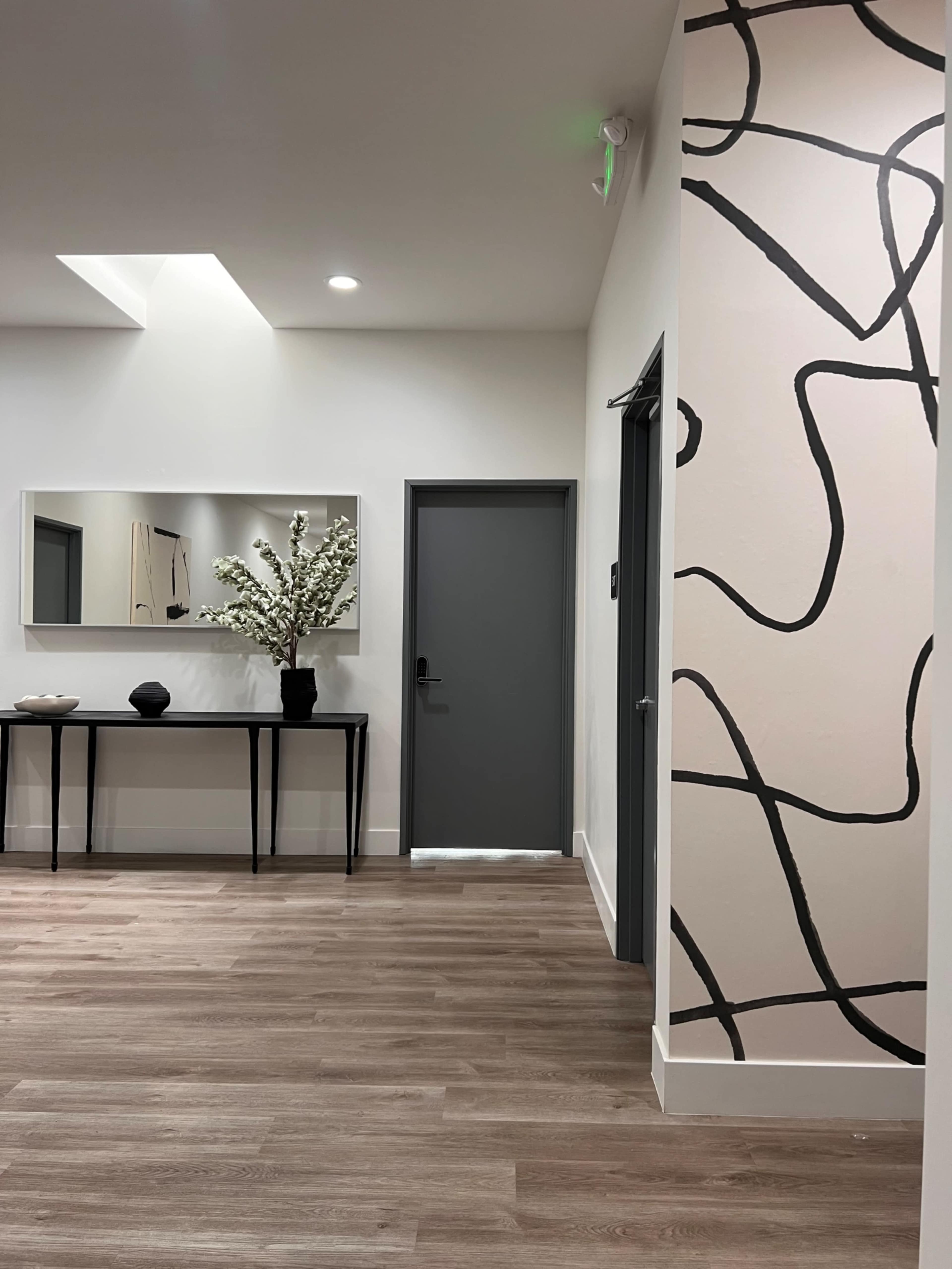 The image shows a modern hallway with a dark door, a large mirror, a console table, and a decorative wall featuring an abstract black line design.
