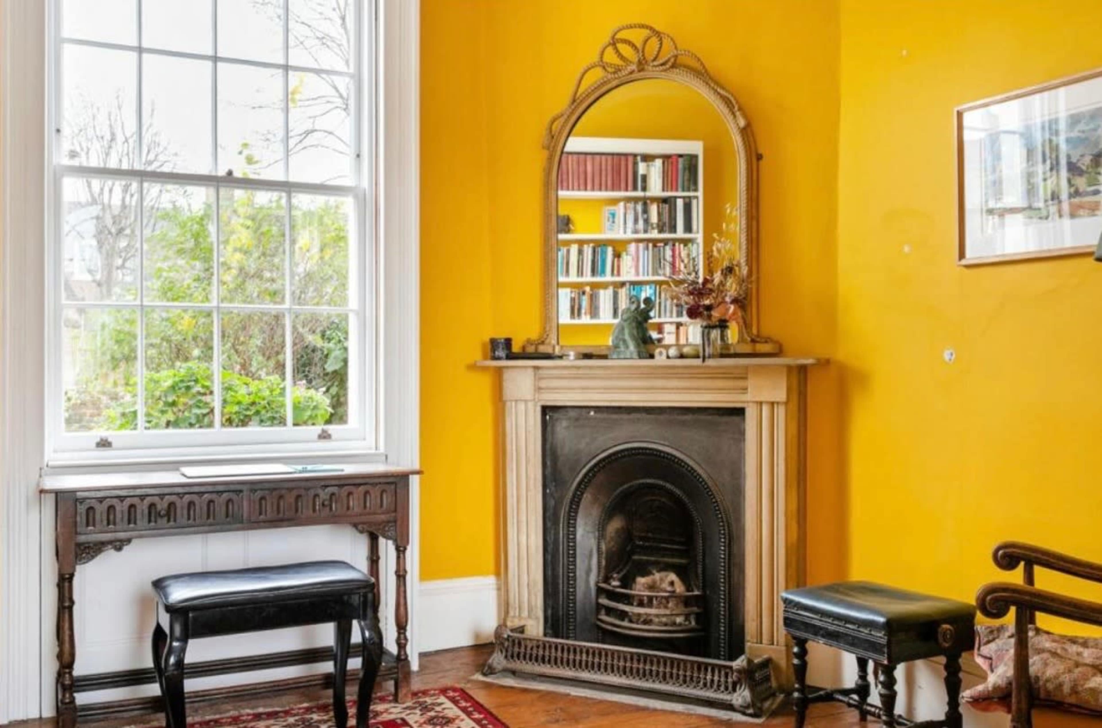 The room features a yellow wall with a fireplace, a large mirror above it, a bookshelf in the background, and two pieces of furniture: a black bench and a chair.