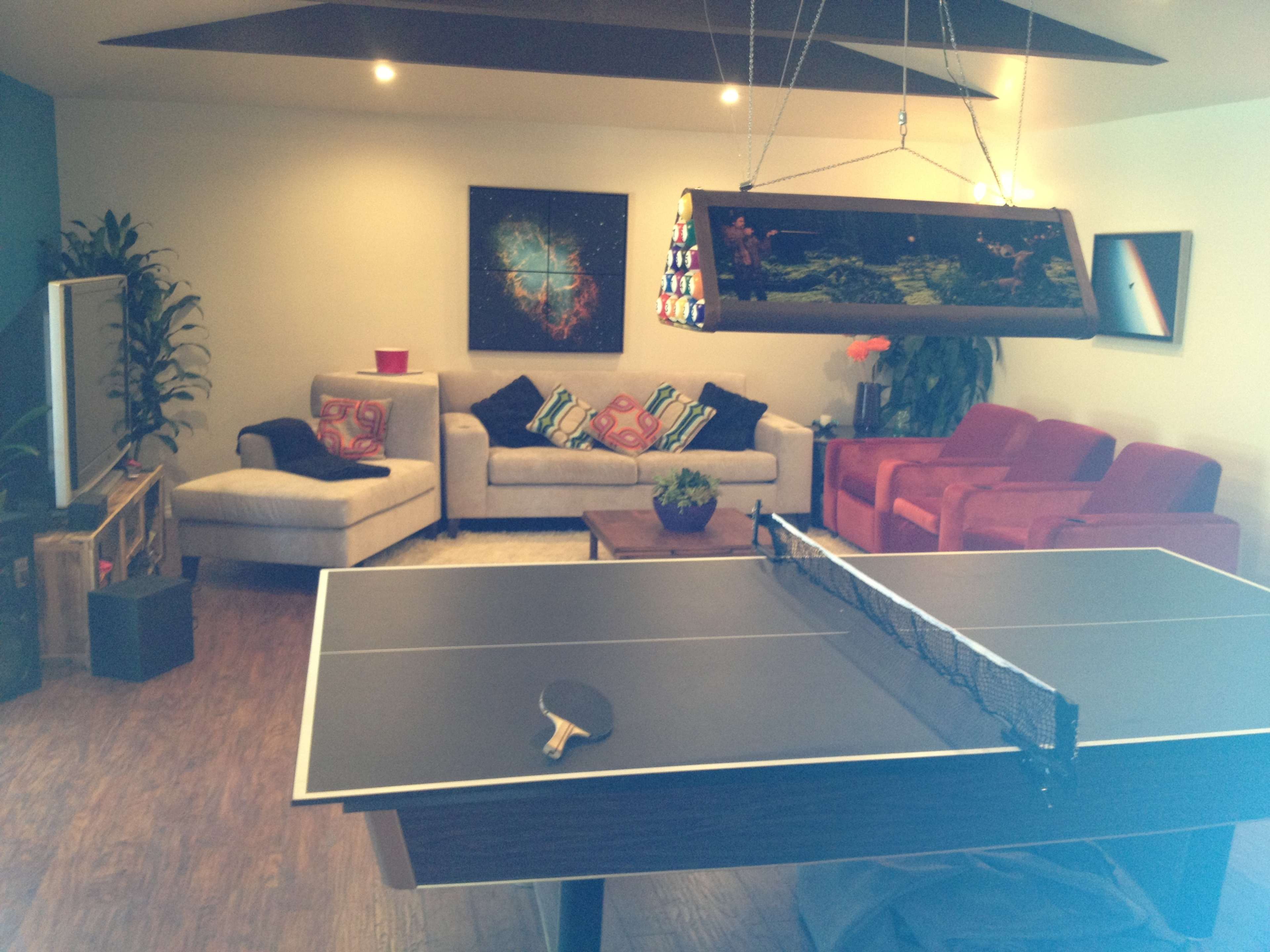 A game room features a ping pong table in the foreground, with a sofa and chairs in the background, along with decorative plants and a large light fixture.