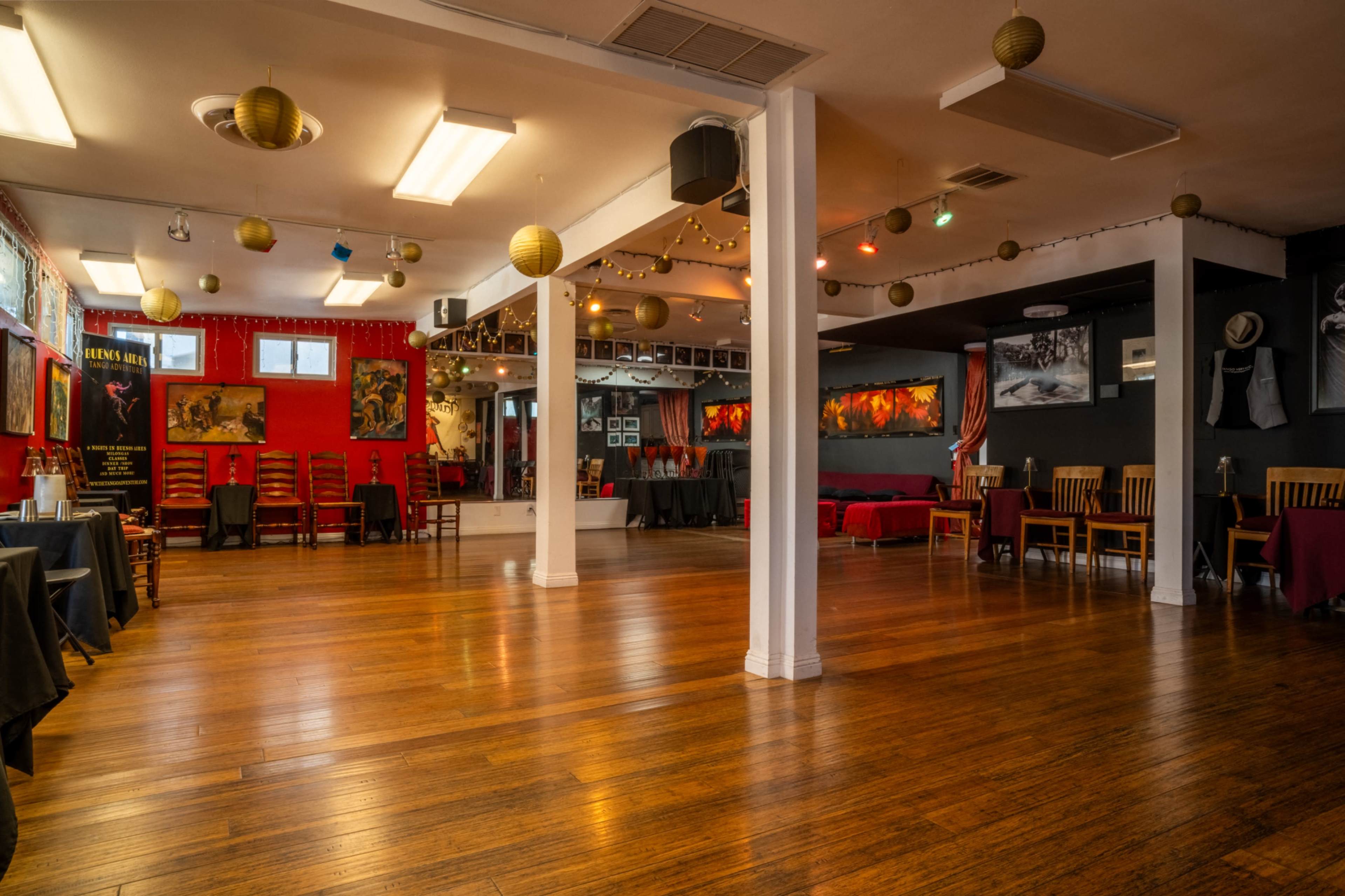 The image shows a spacious dance studio with wooden floors, red and black walls adorned with artwork, and a few scattered chairs and tables.